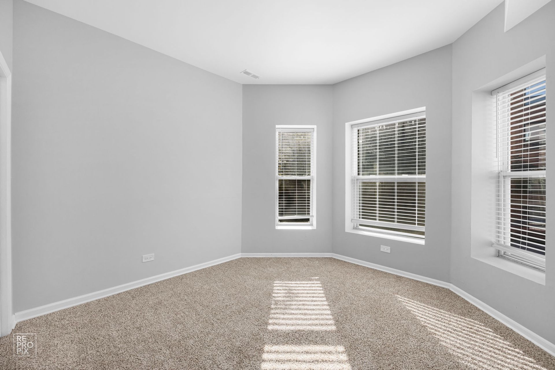 Empty room with light gray walls, carpeted floor, and three windows with blinds.