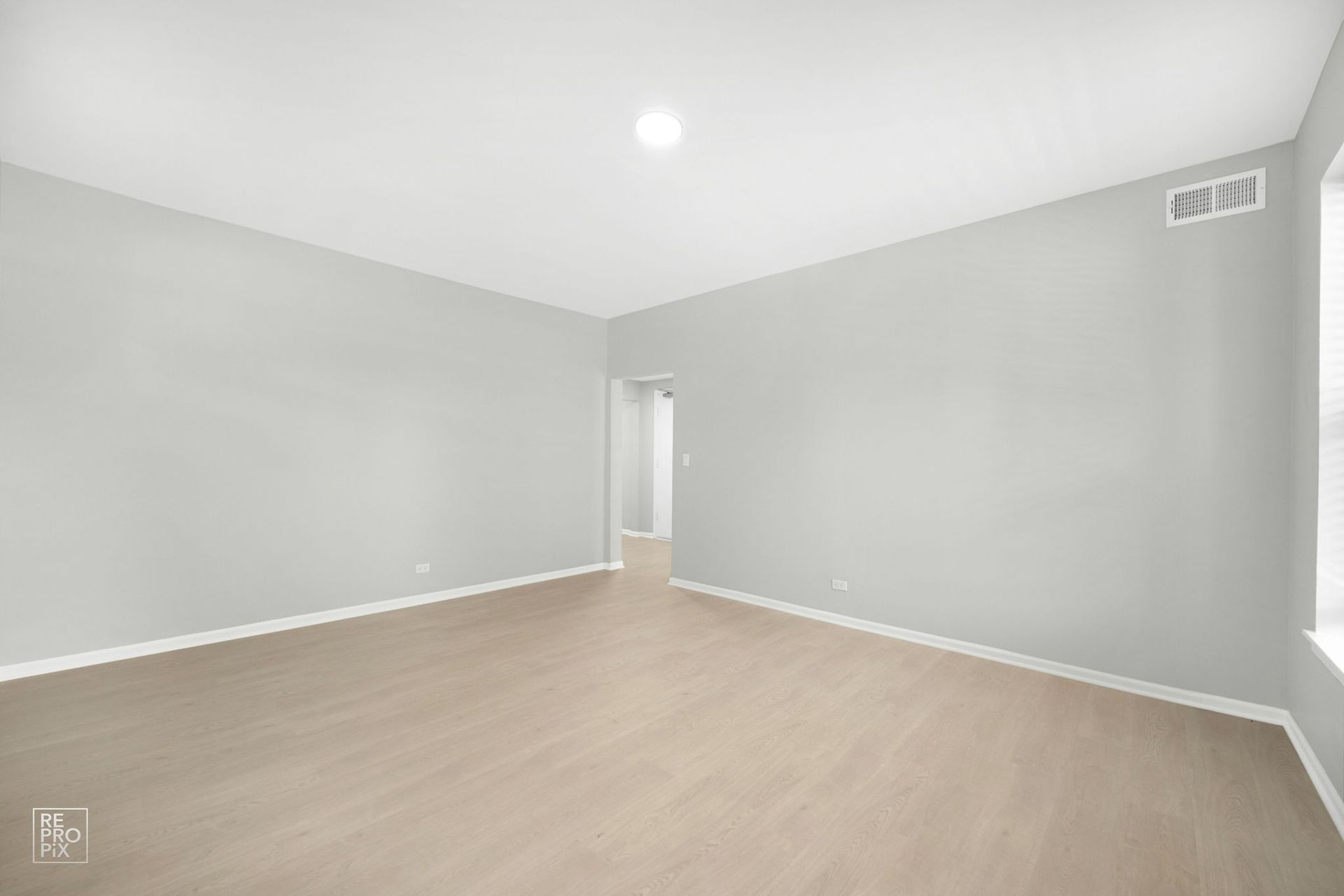 Empty room with gray walls and light-colored flooring. A doorway leads to another room.