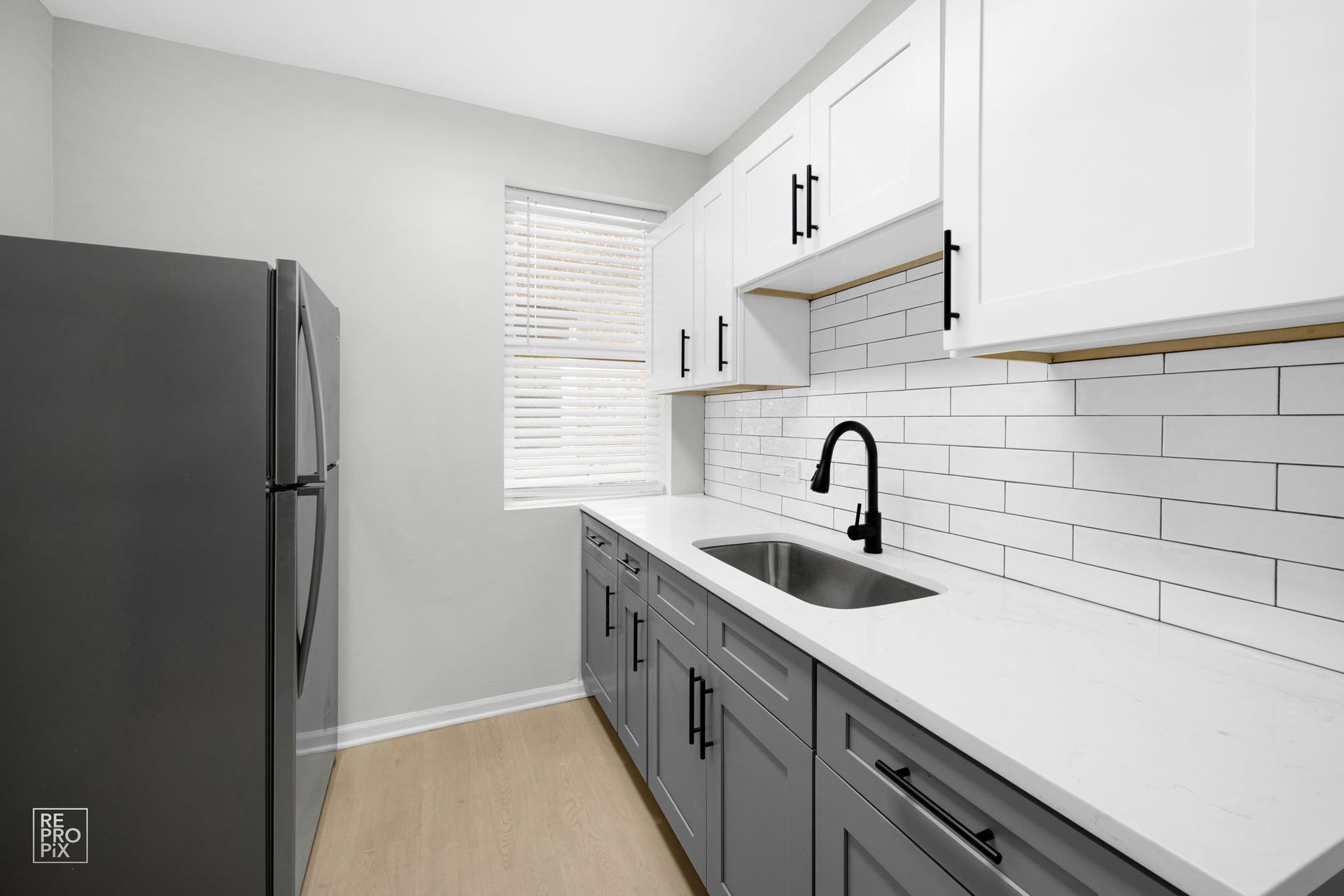 Modern kitchen with gray and white cabinets, stainless steel sink, subway tile backsplash, and light wood floor.