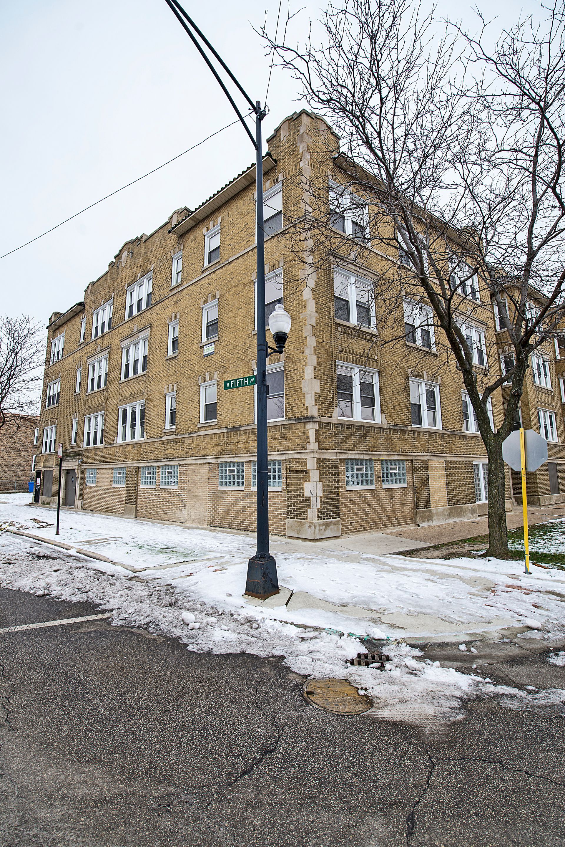 Multi-story brick building on a snowy street, with a street lamp in front and bare trees.