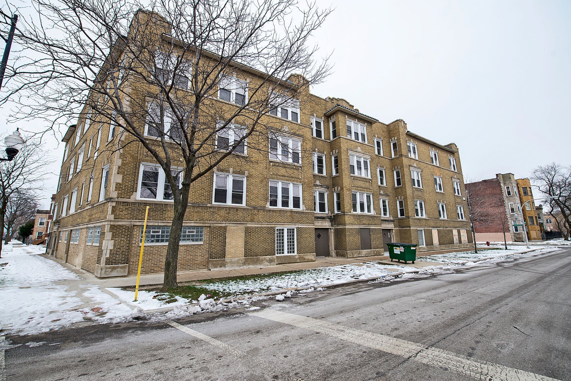Stone apartment building on a snowy street. Bare tree in front, overcast sky.