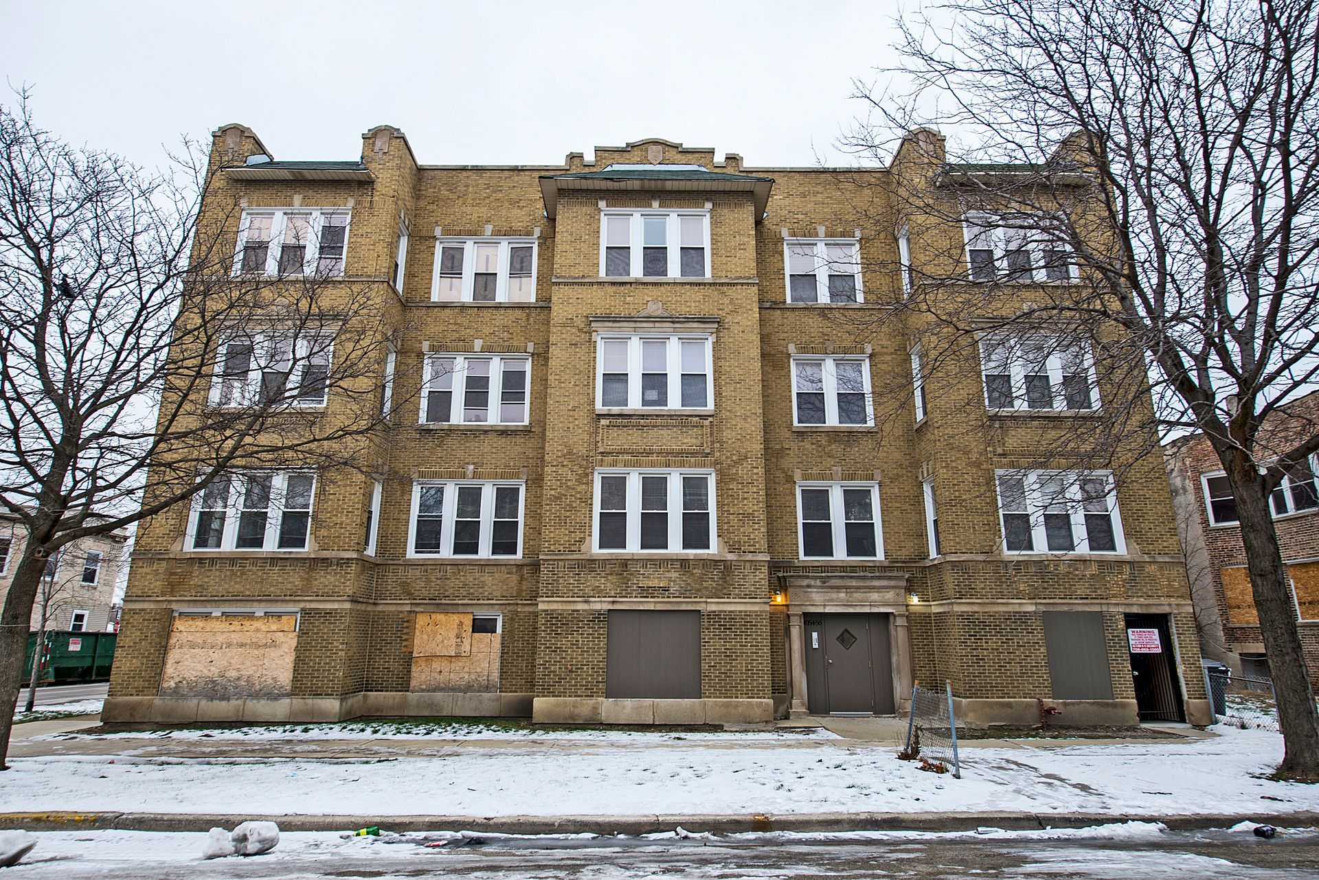 Multi-story brick apartment building on snowy street; boarded-up windows, leafless trees.