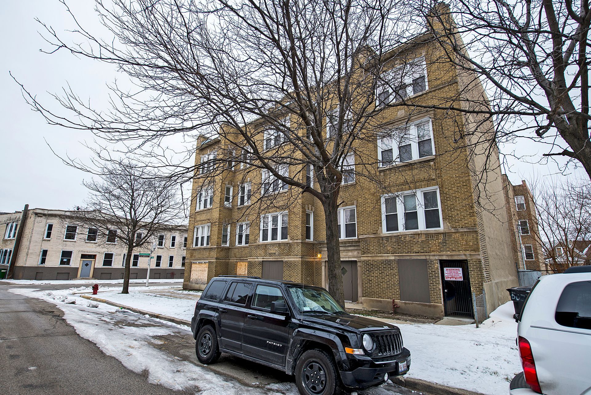 Apartment building with boarded-up windows and snow-covered ground. Black SUV parked on street. Bare trees and overcast sky.