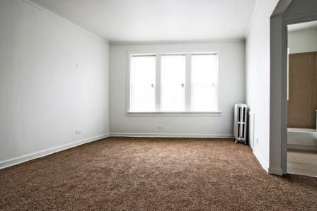 Empty room with brown carpet, three-pane window, and radiator. Opening to another room on the right.