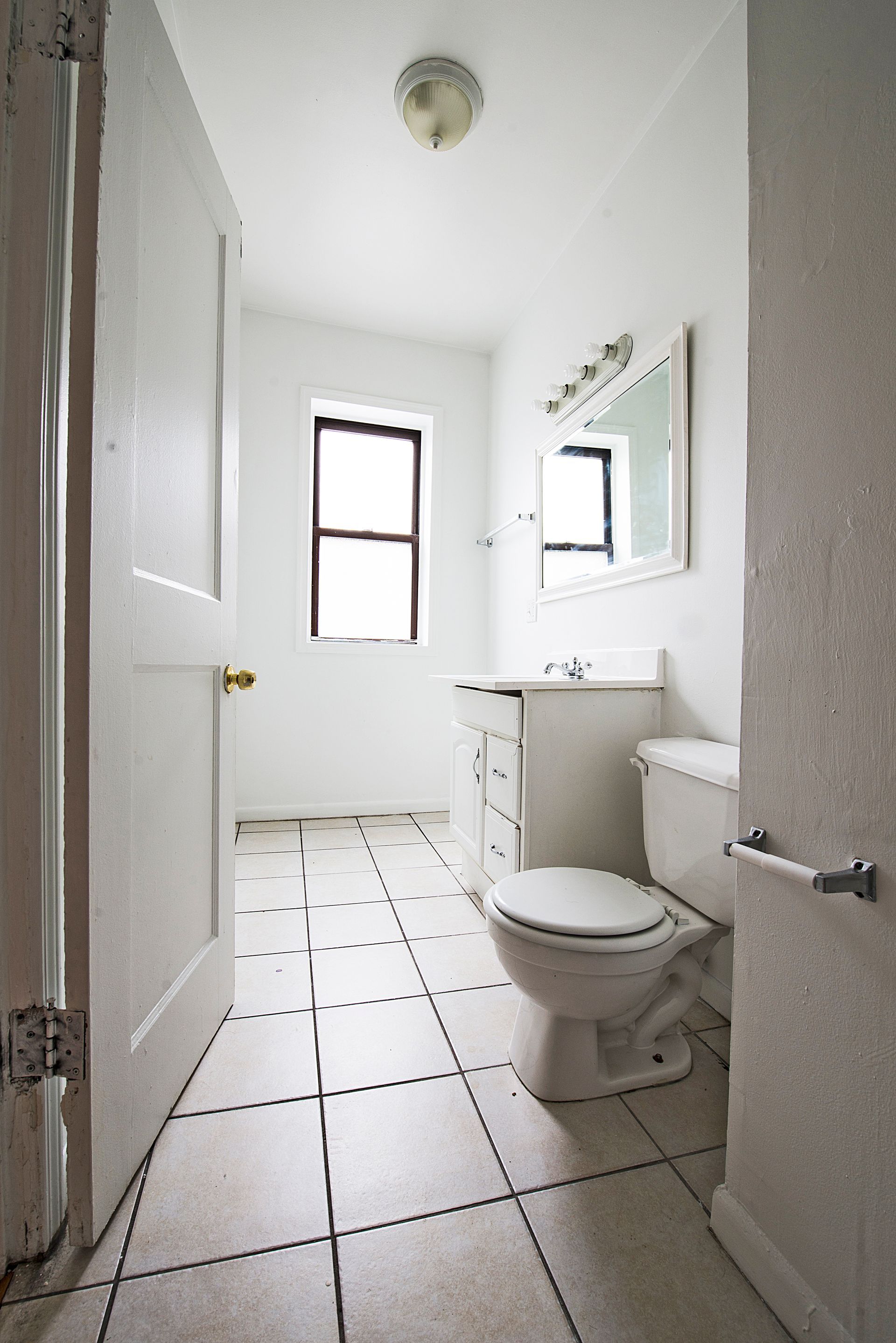 White bathroom with toilet, sink, mirror, window, and tiled floor.