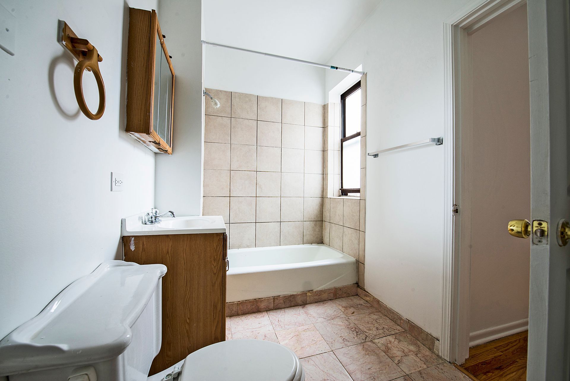 Bathroom with a white toilet, vanity, and tub; beige tile, a small window, and open door.