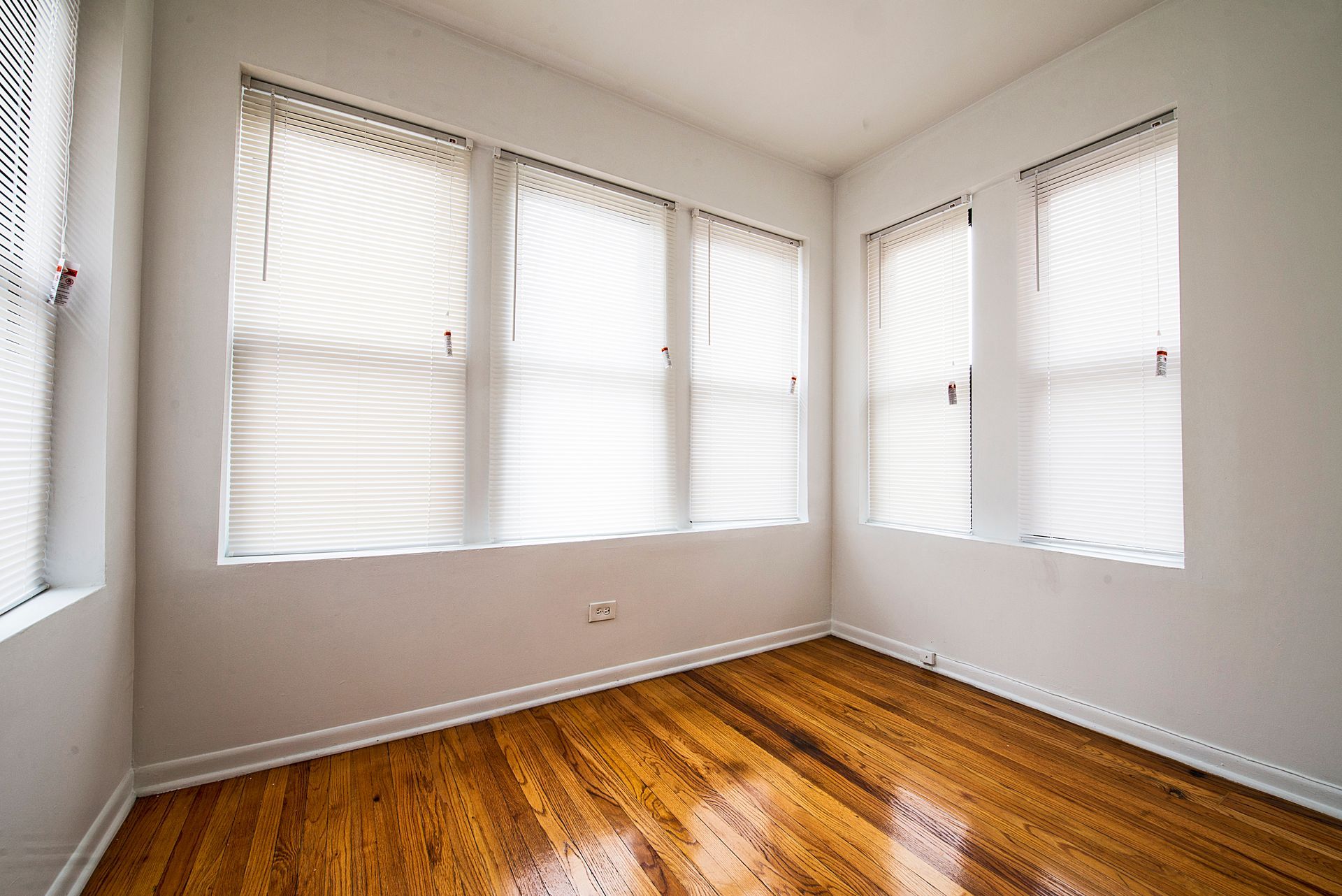 Empty room with hardwood floors, white walls, and three windows with blinds.
