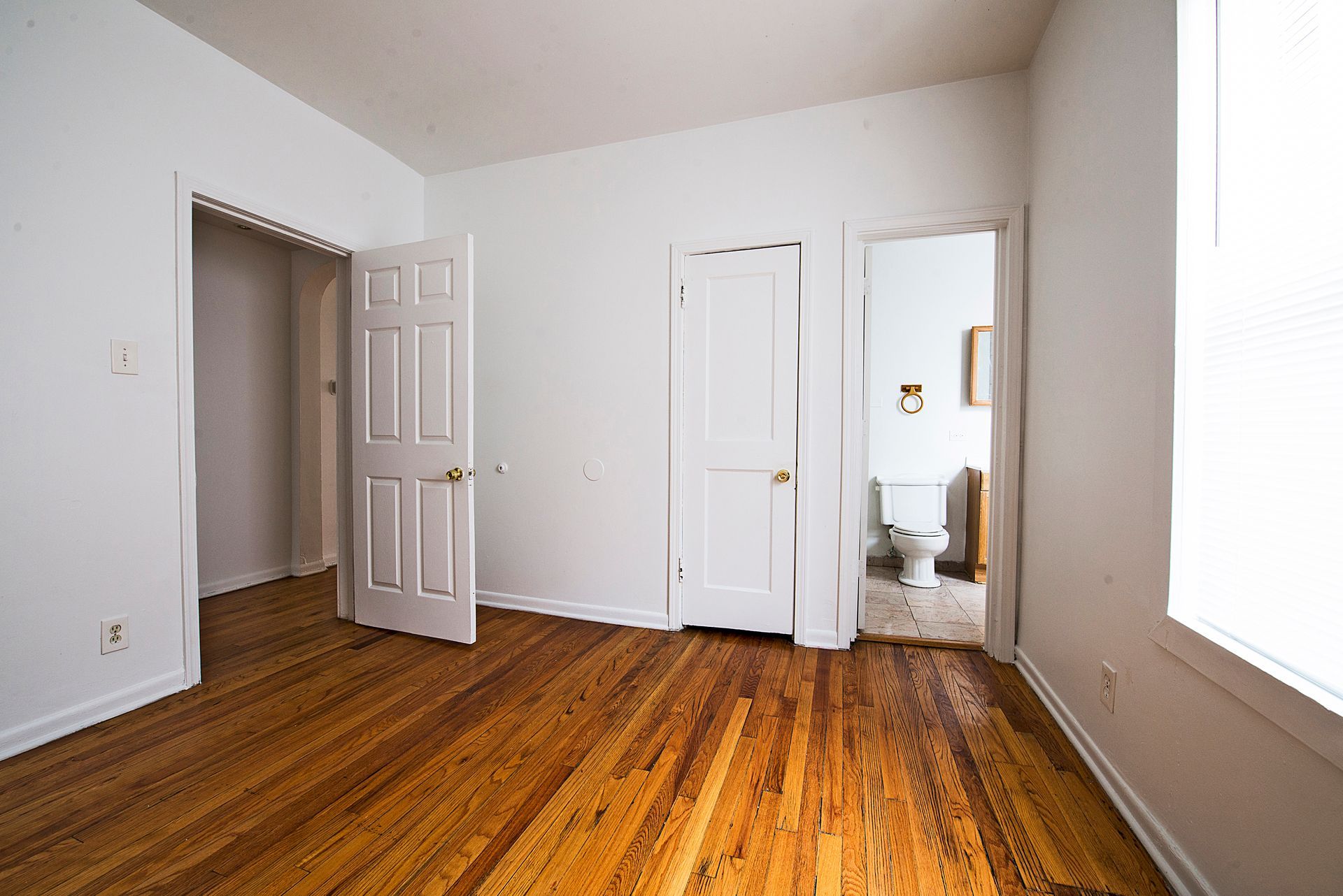 Empty room with wood floor, white walls, and three doorways: closet, bedroom, and bathroom.
