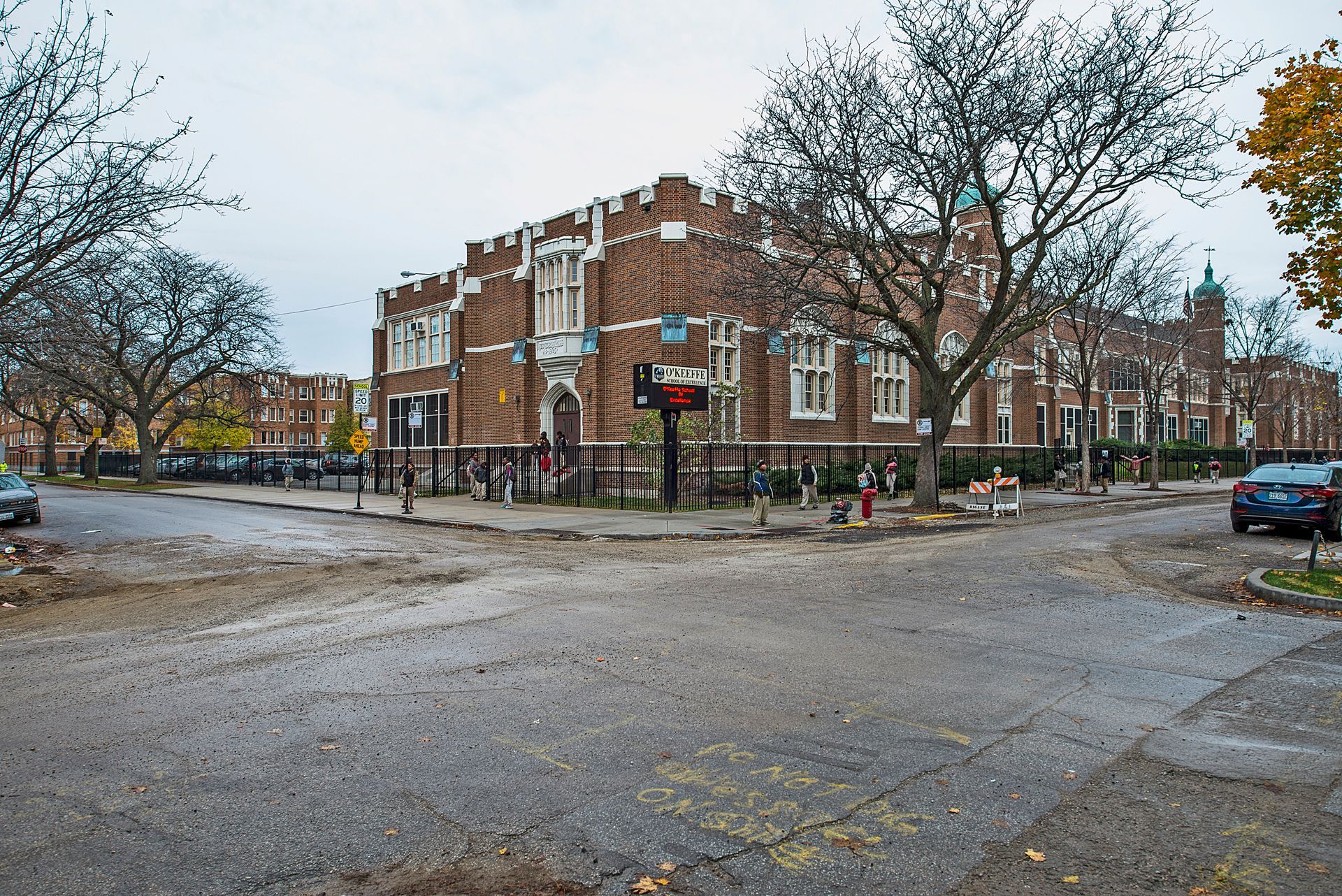 Brick building on a corner with trees. Gray, overcast sky. Cars parked on streets.