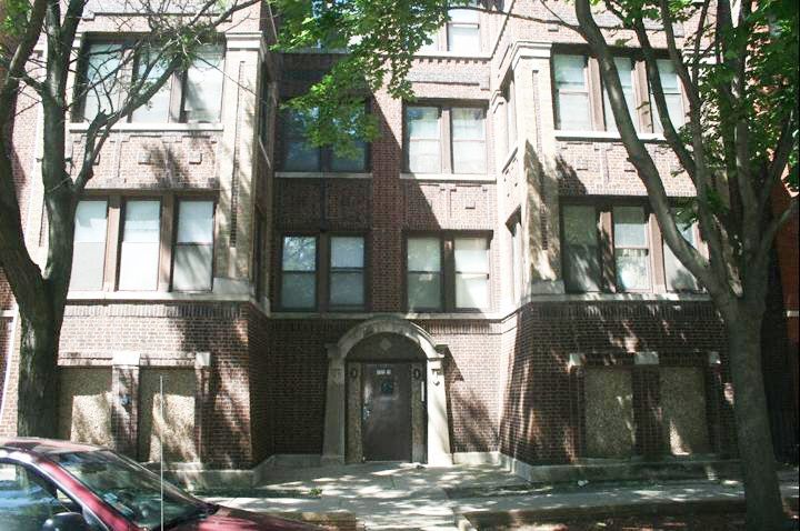Brick apartment building facade with a central arched entrance and windows. Shade from trees.