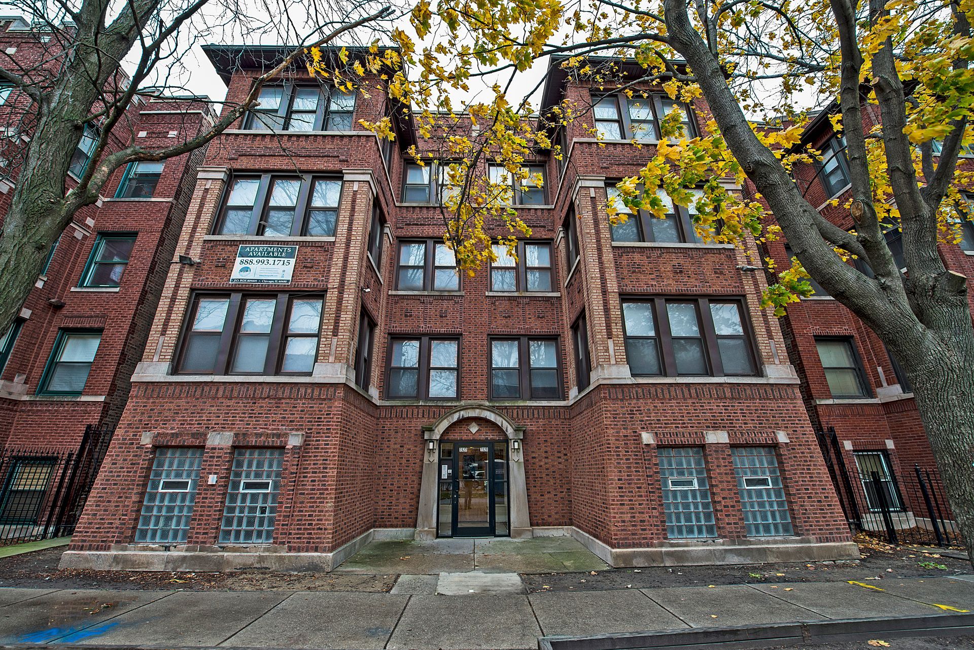 Brick apartment building with multiple stories and windows, trees with yellow leaves in the foreground.
