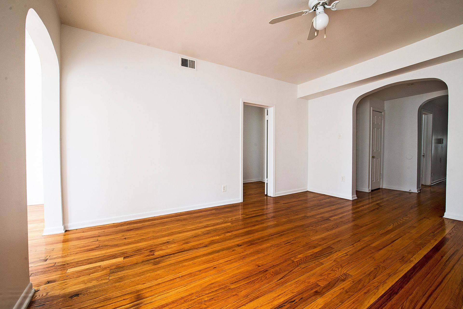 Empty room with hardwood floors, white walls, arched doorways, and a ceiling fan.