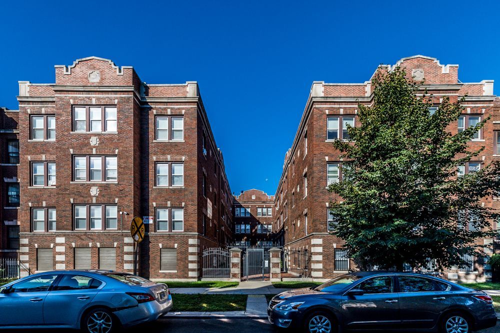 Two matching brick apartment buildings with cars parked in front under a blue sky.