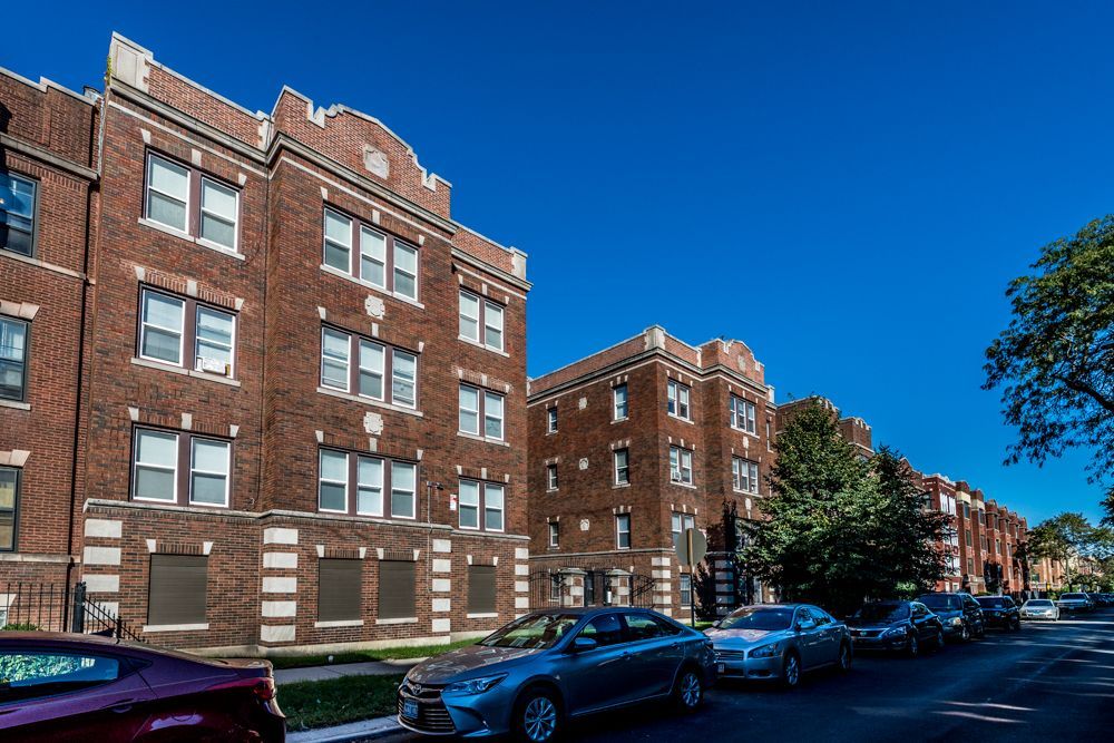 Brick apartment buildings on a city street with parked cars and a clear blue sky.