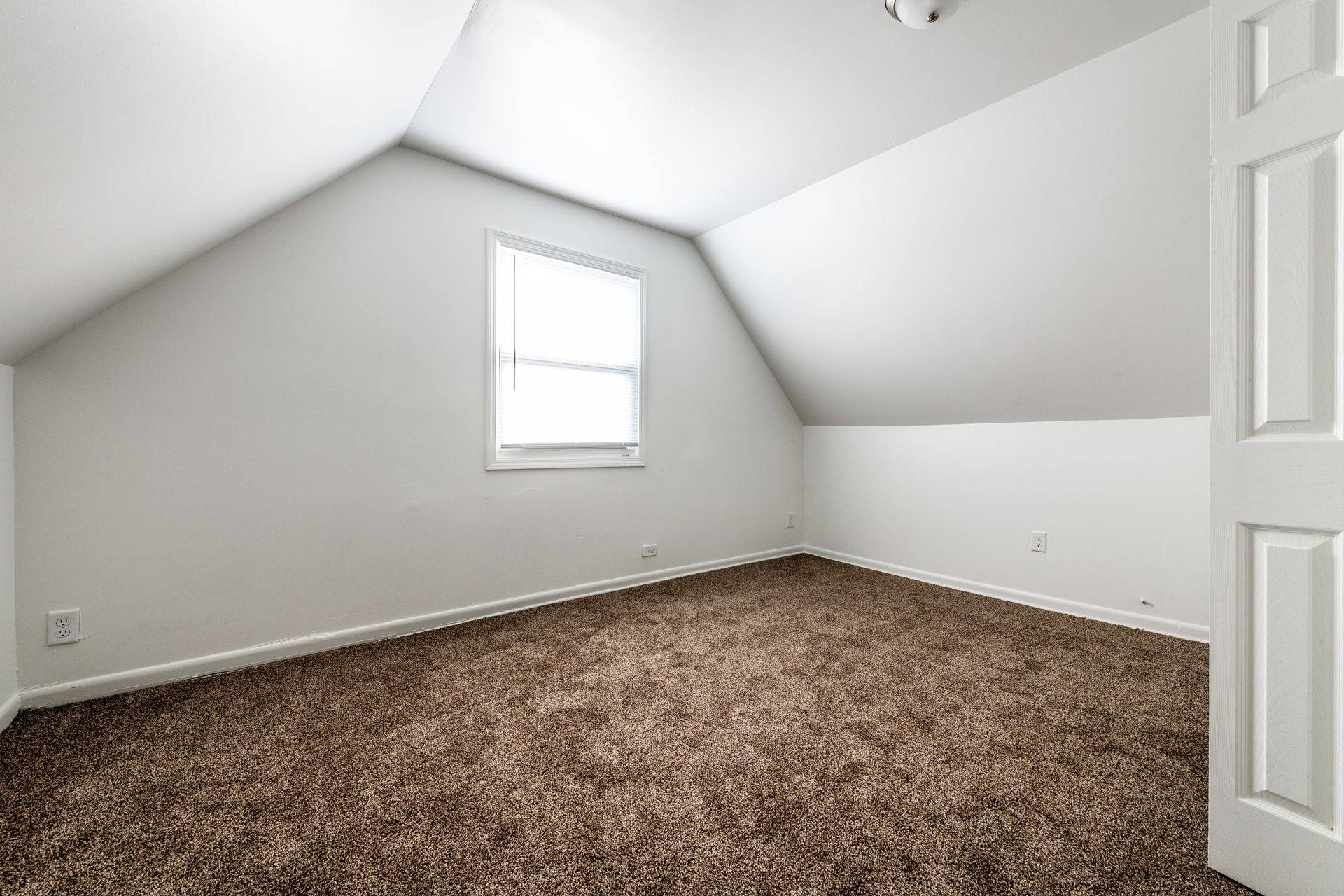 Empty attic room with brown carpet, white walls, window.