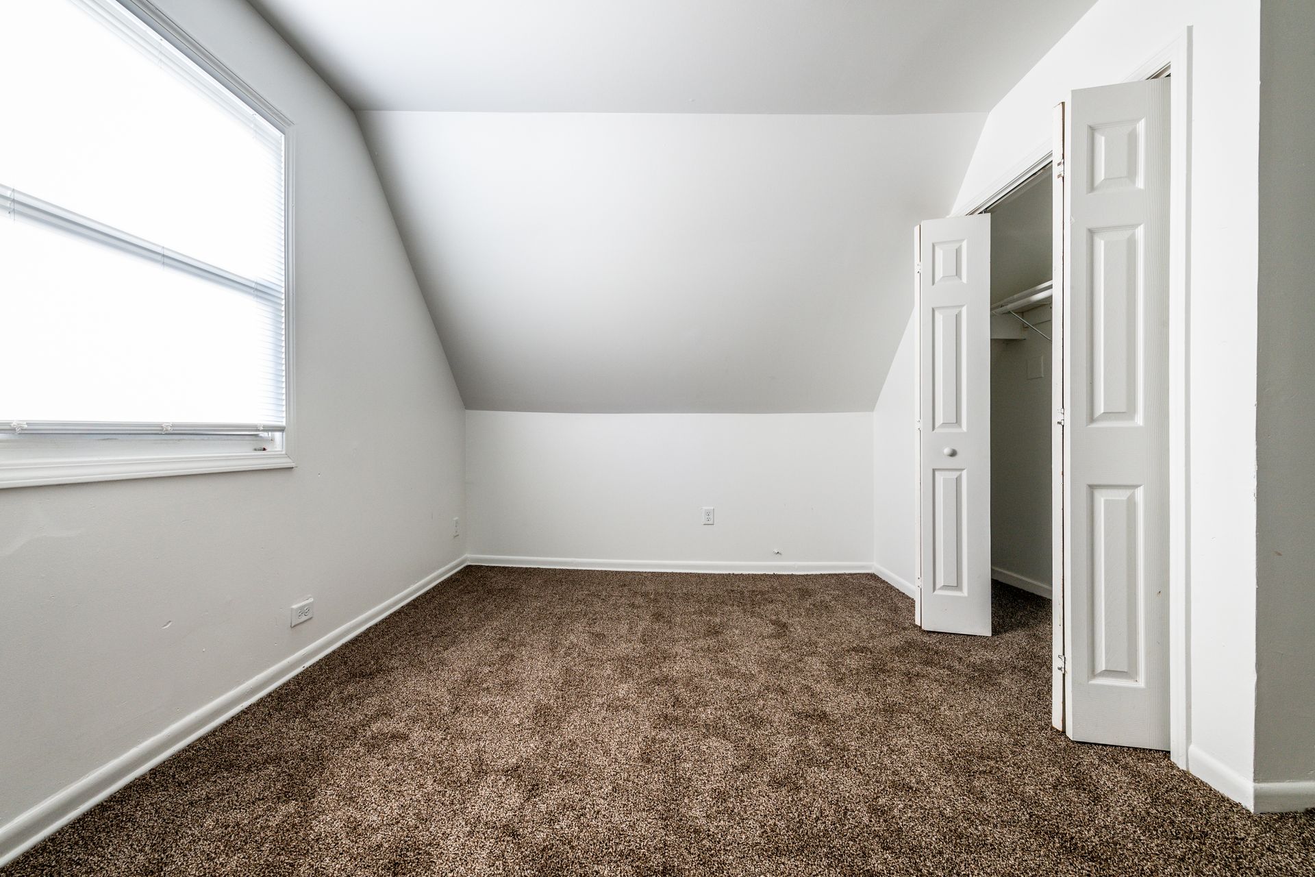 Empty room with brown carpet, sloped ceiling, window, and closet with open doors.