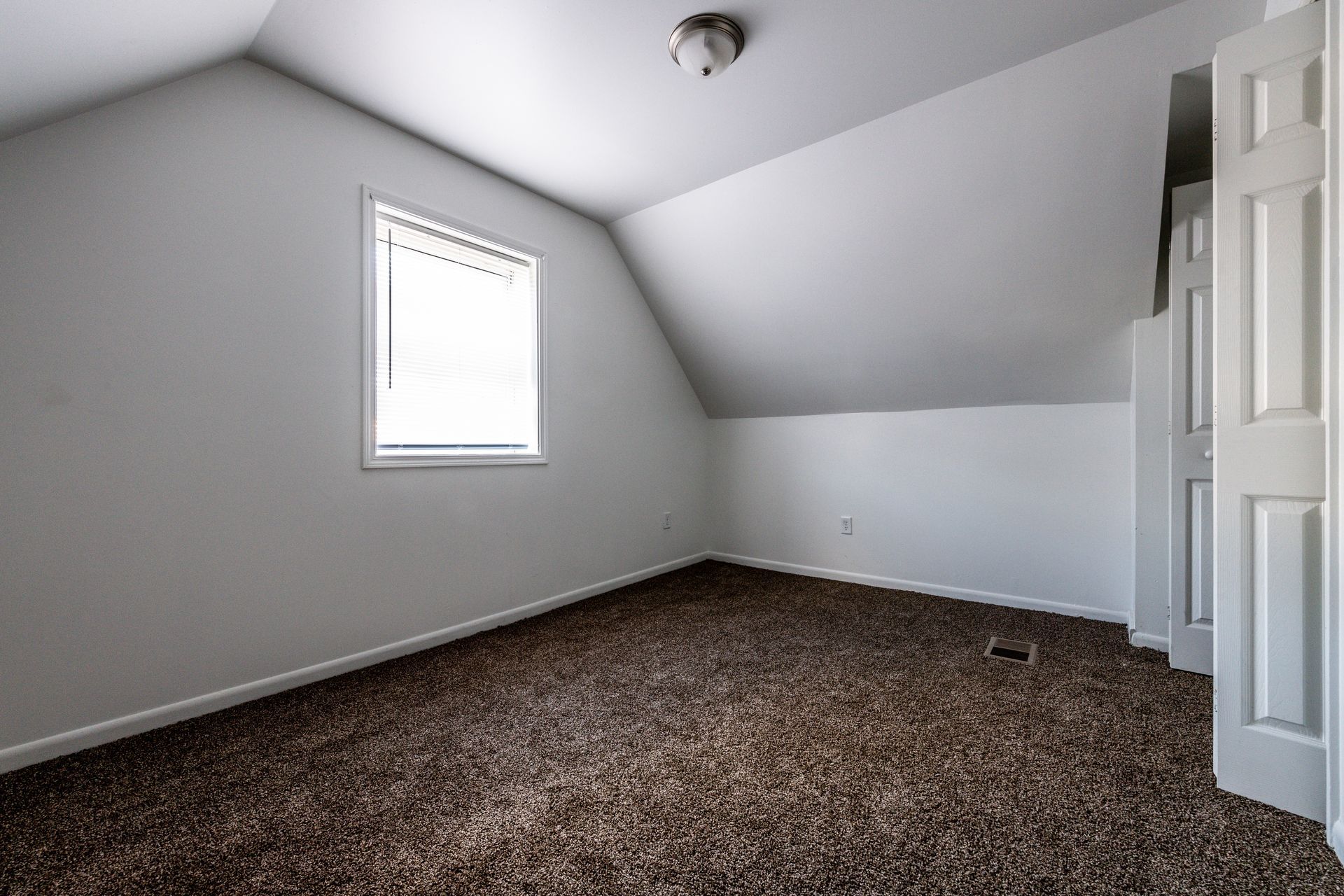 Empty room with brown carpet, white walls, angled ceiling, small window, and closet.