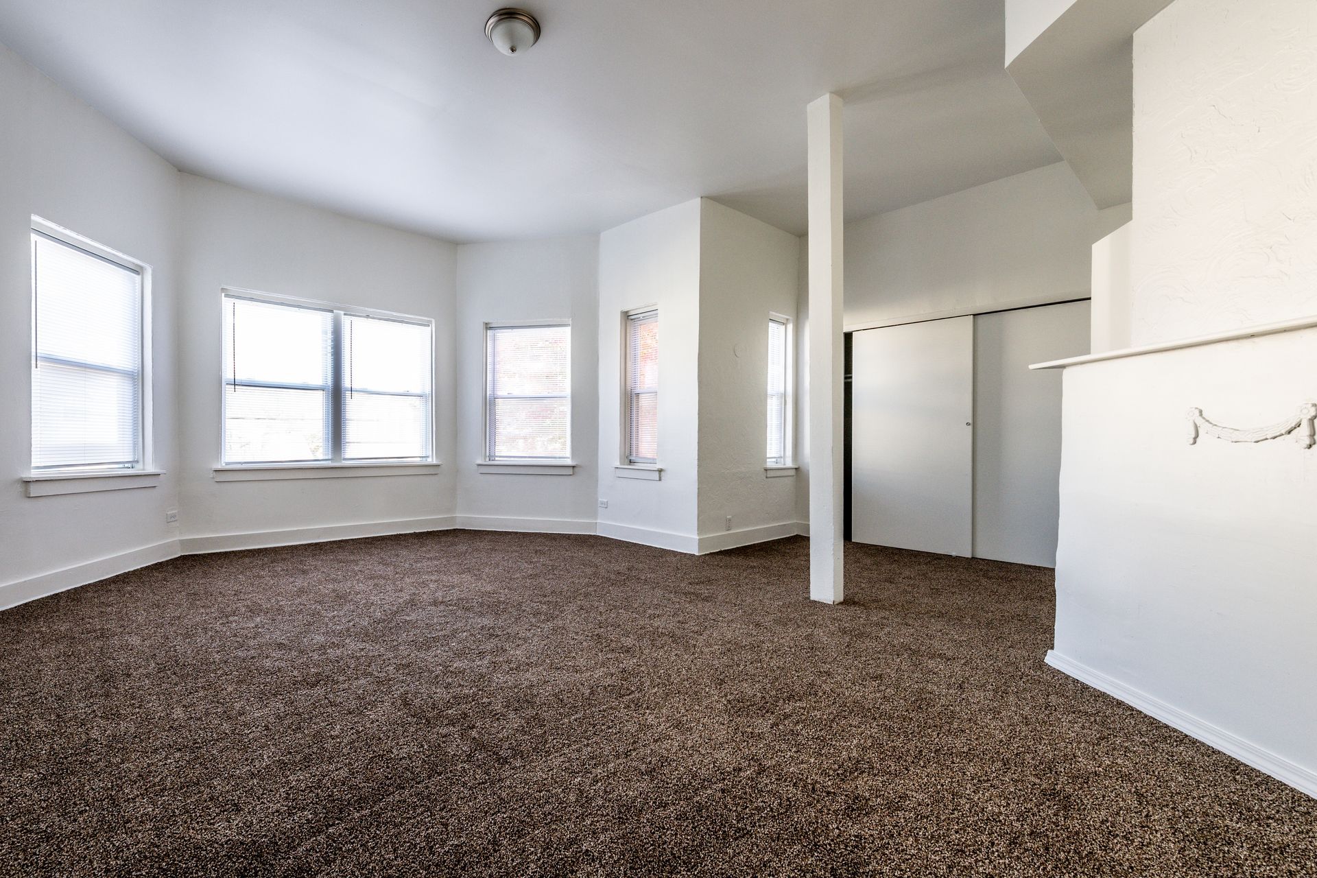 Empty room with brown carpet, white walls, and a column. Bay windows on the left.