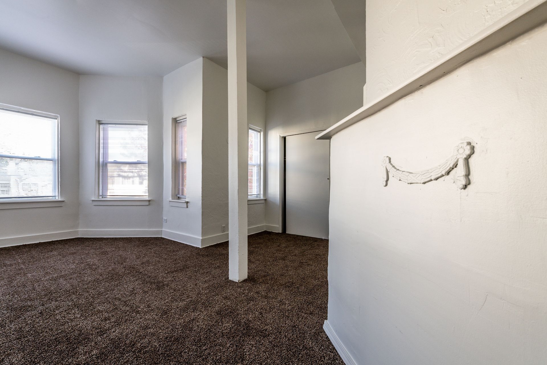 Empty room with brown carpet, white walls, and three windows. A pillar stands in the center.