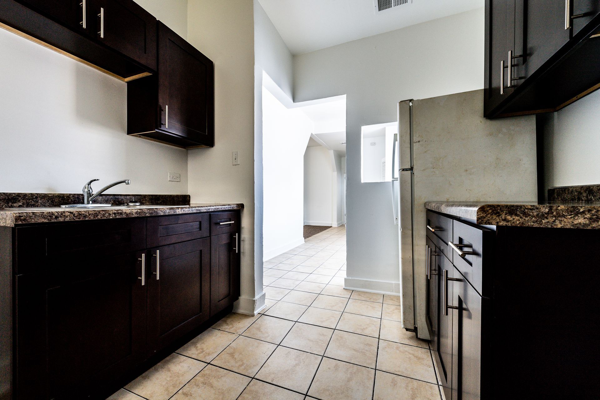 Kitchen with dark cabinets, white walls, and a light-colored tile floor. Refrigerator visible.