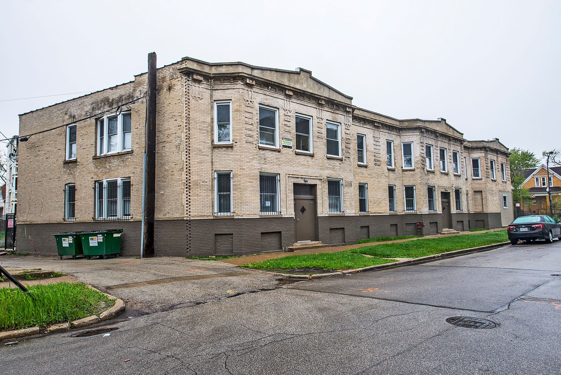 Two-story brick apartment building on a corner lot, green trash bins, overcast sky, street view.