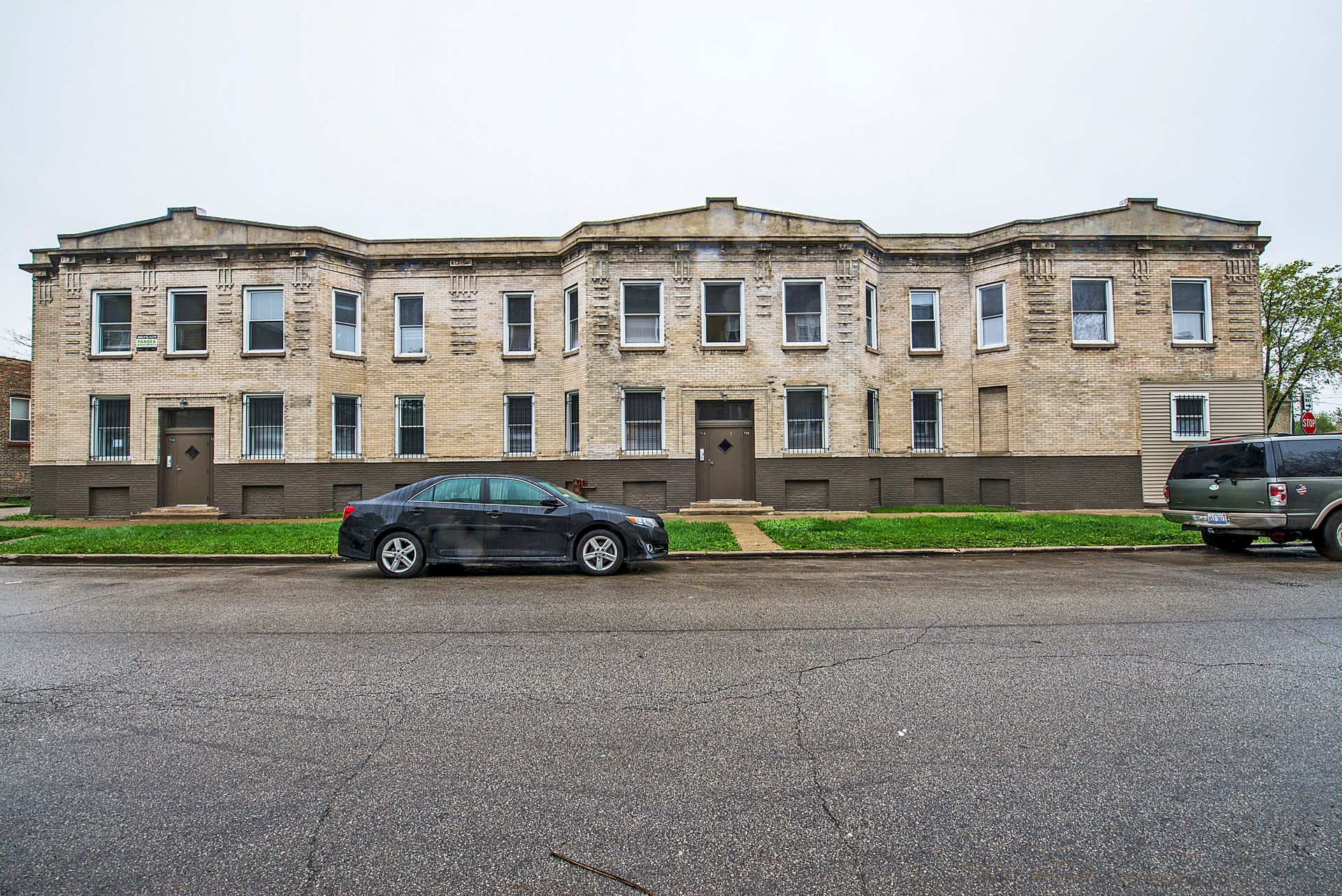 Gray two-story apartment building with stone facade; parked cars on wet street.