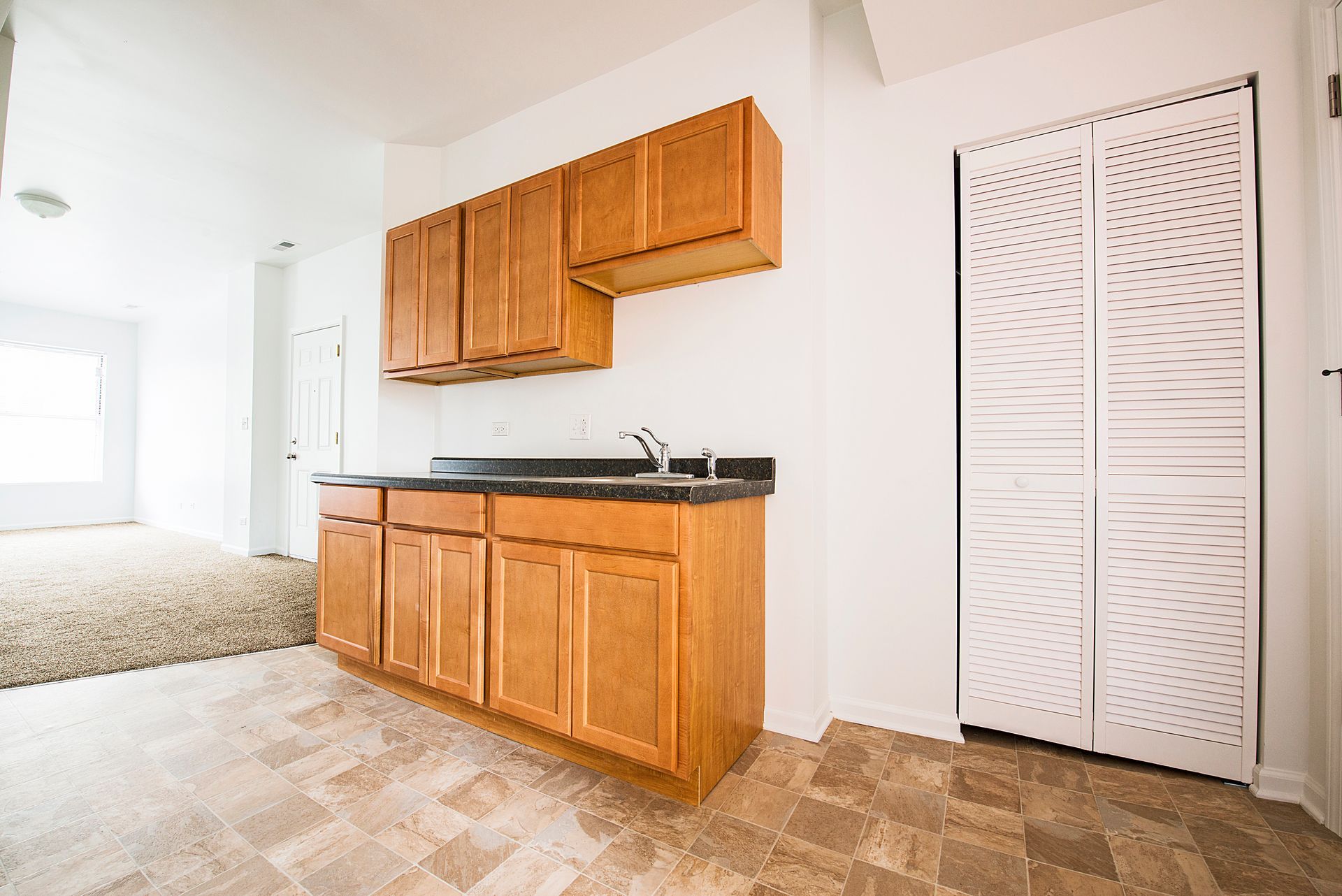 Small kitchenette with wood cabinets, countertop, and sink. White folding door to the right. Beige flooring.