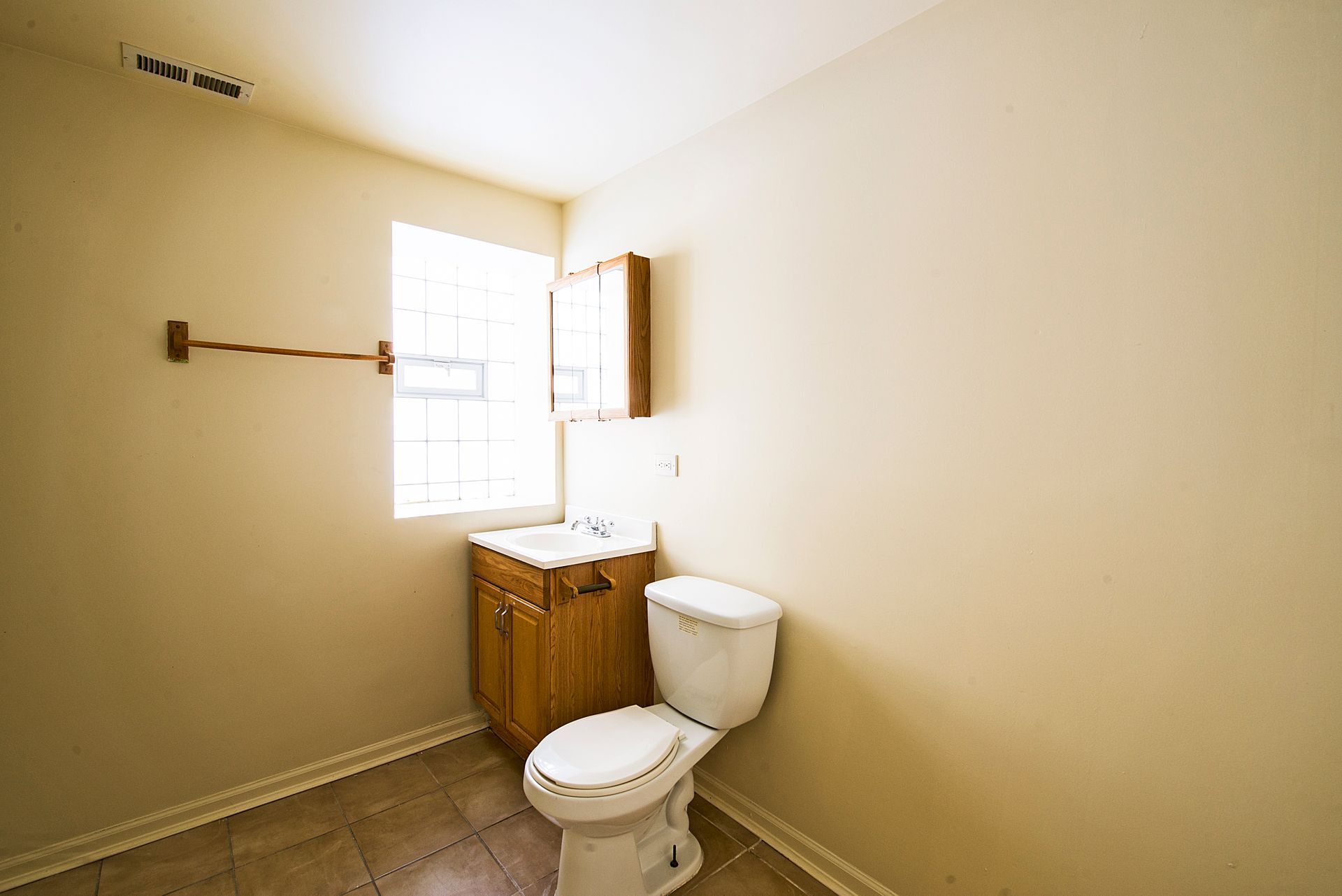 Bathroom with toilet, sink in a wooden cabinet, window, and medicine cabinet. Beige walls, tile floor.