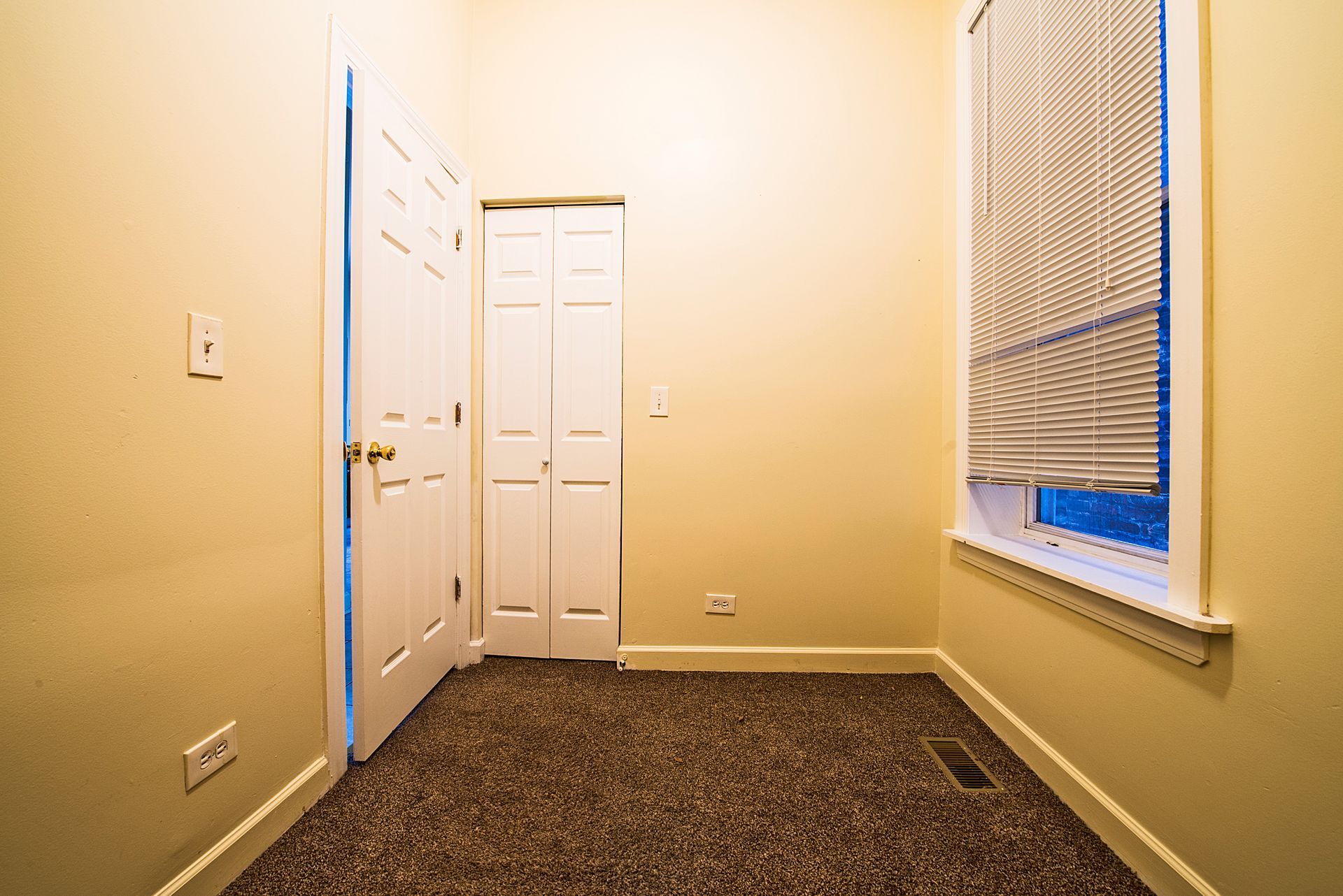 Empty room with beige walls, two white doors, carpet, and a window with blinds.