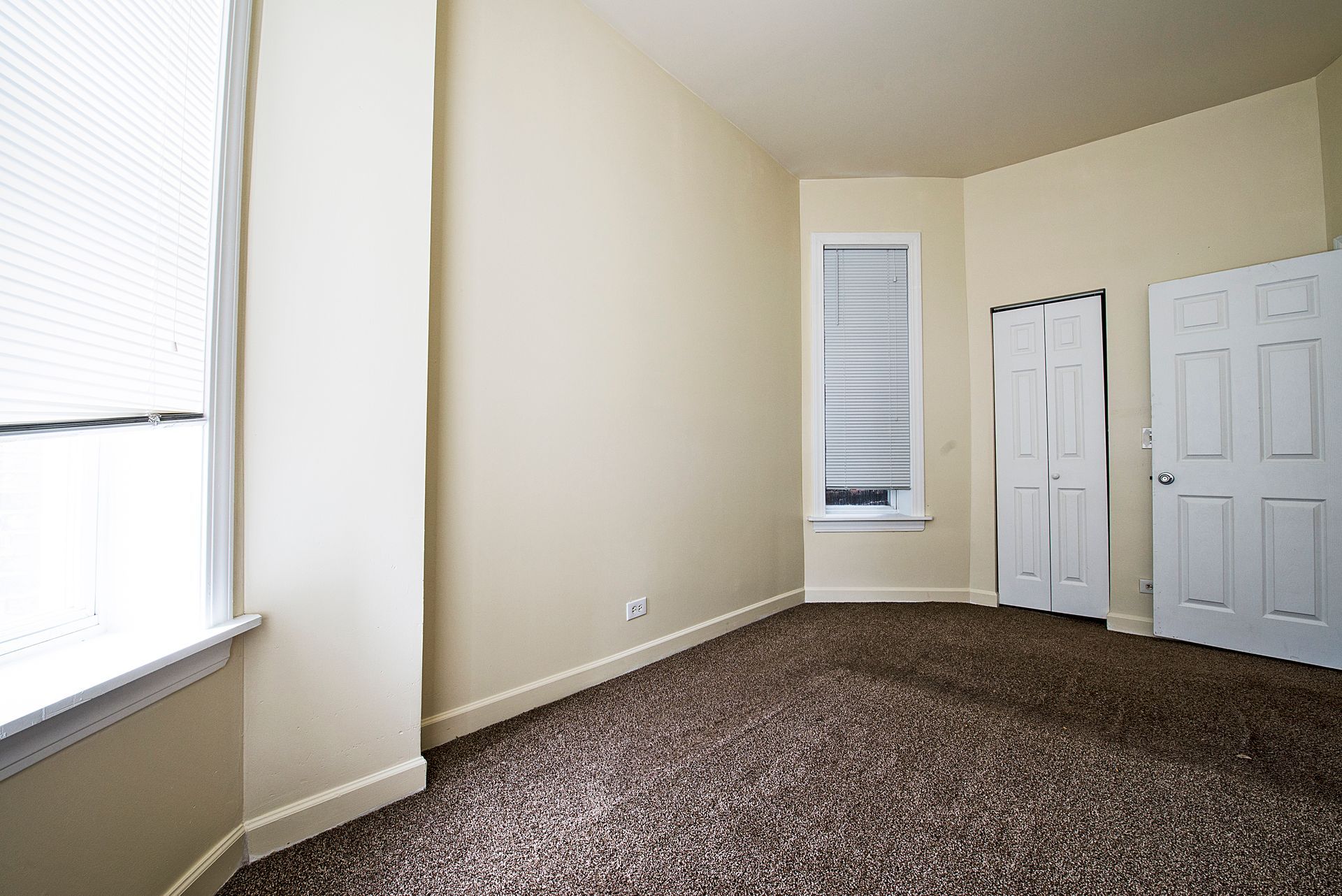 Empty room with tan walls, brown carpet, two windows, and two white doors.