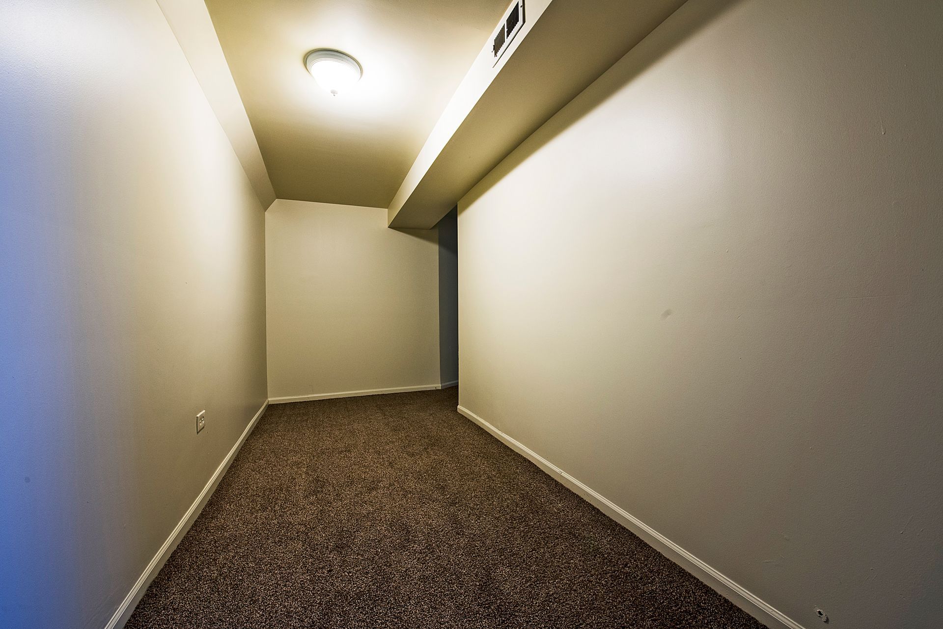 Small, empty room with beige walls and brown carpet. Bright overhead light.