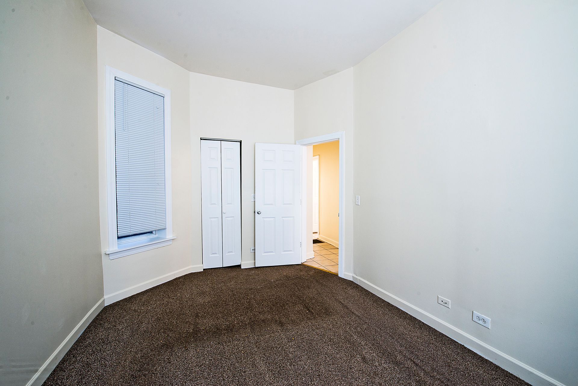 Empty bedroom with brown carpet, white walls, closet, and a window with blinds.