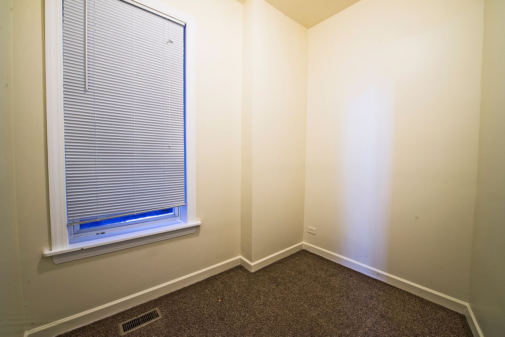 Empty room with a window covered by blinds; beige walls, brown carpet, and a white trim.