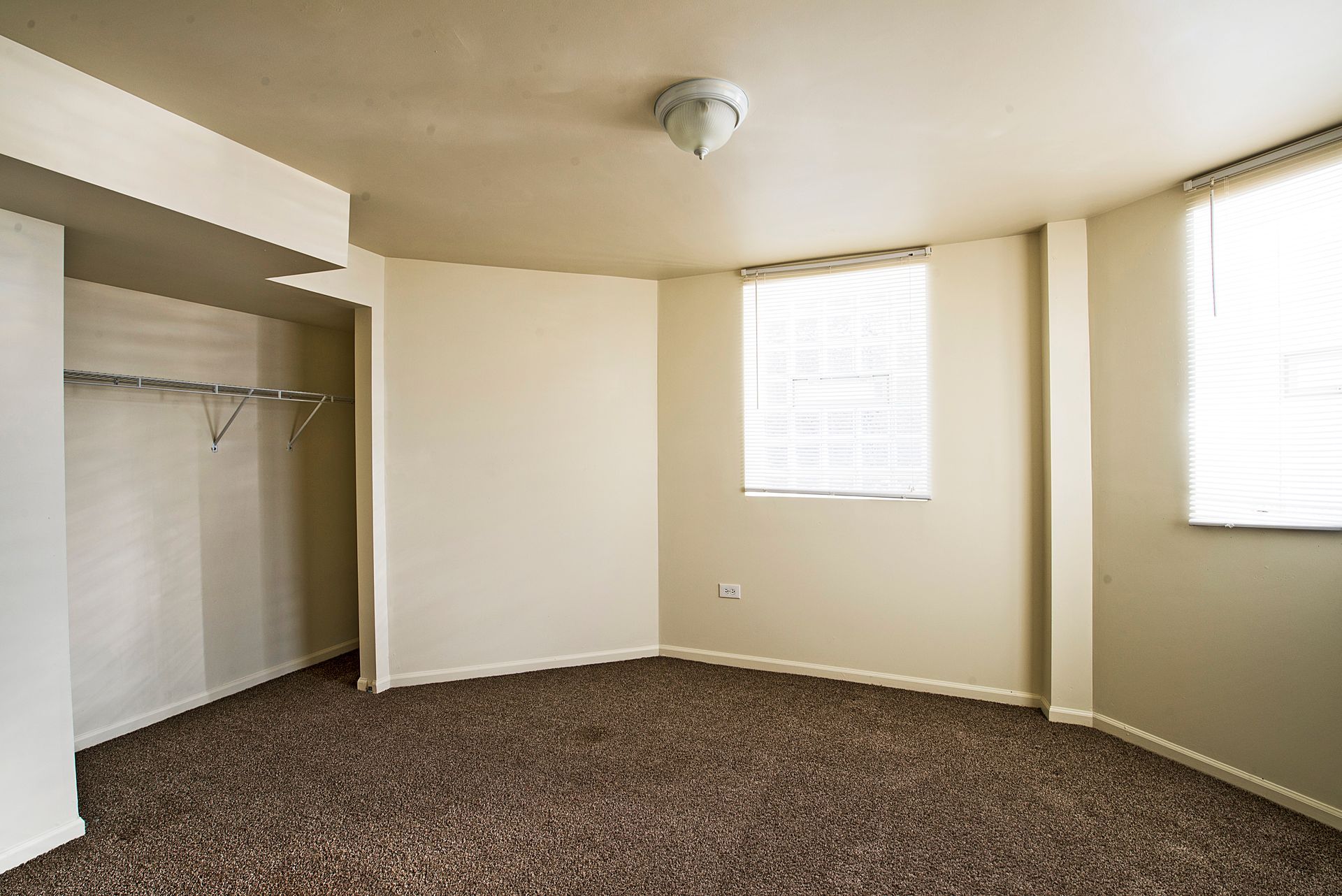 Empty bedroom with brown carpet, closet, two windows, and overhead light.