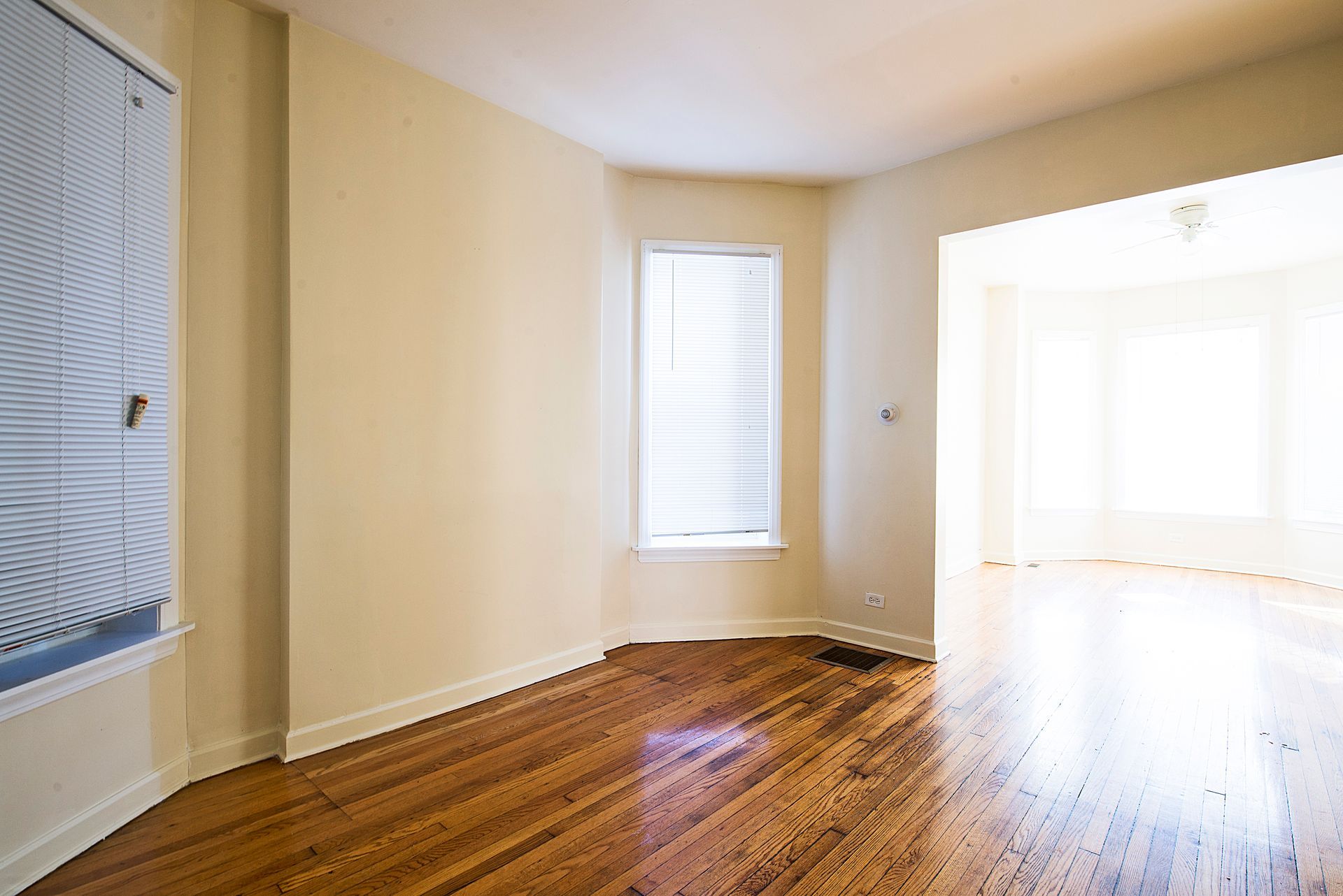 Empty room with hardwood floors, a window with blinds, and an open doorway leading to a bay window.