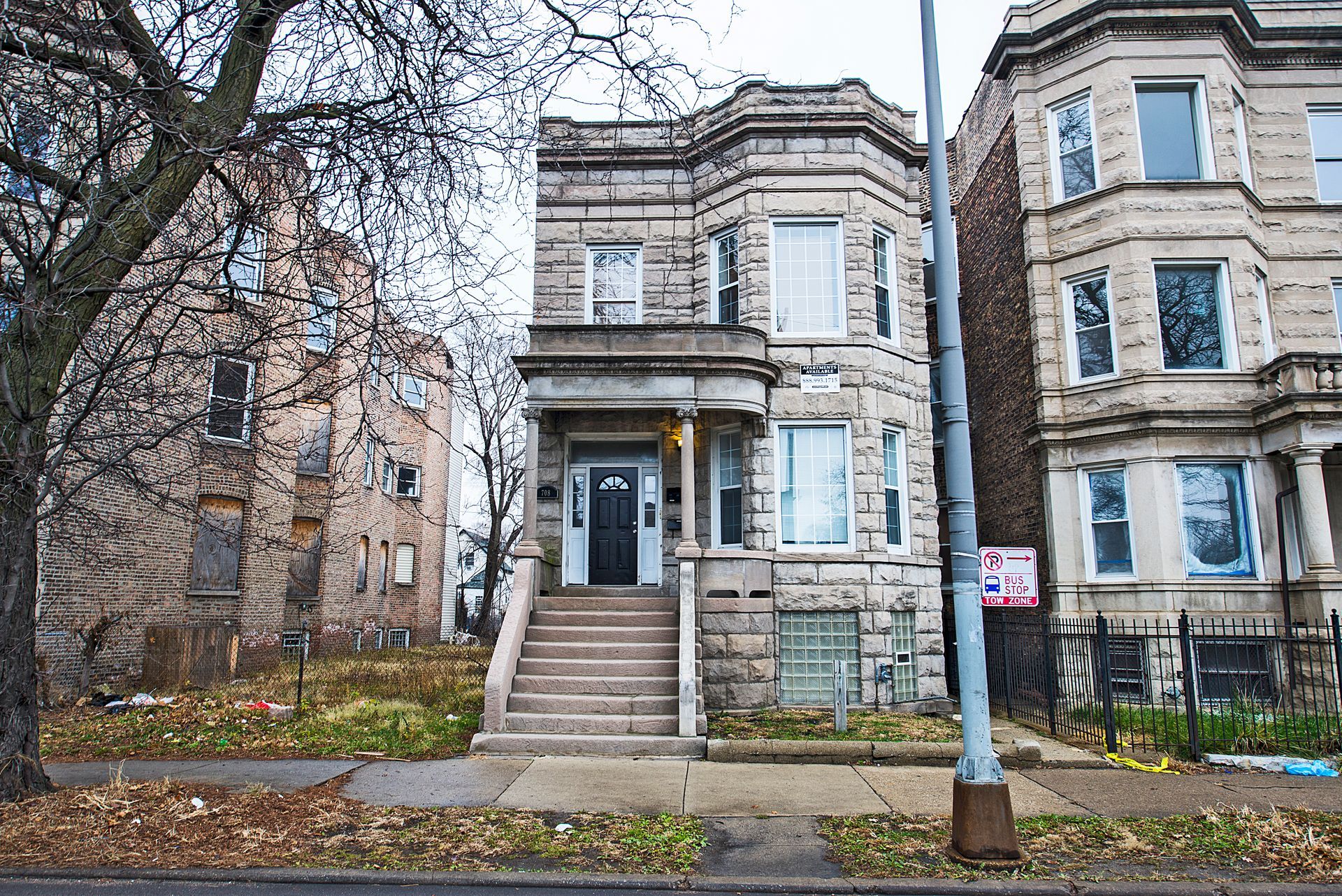 Three-story stone building with porch and stairs, flanked by other buildings in a residential area.