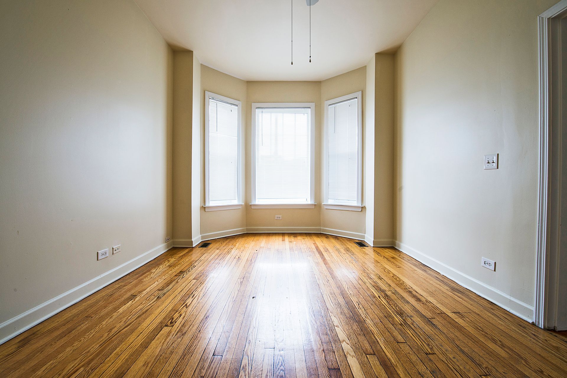 Empty room with hardwood floors, a bay window with curtains, and off-white walls.