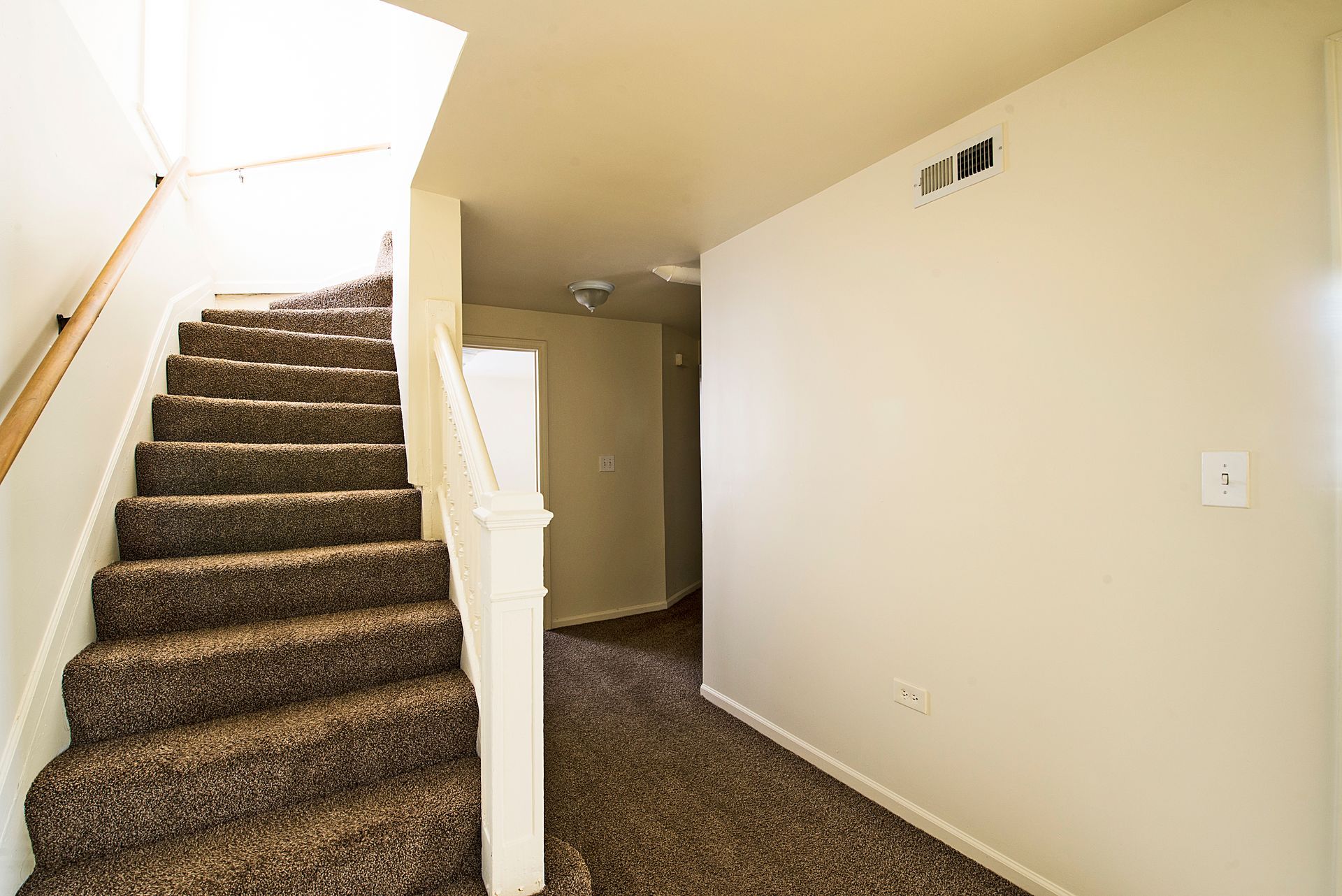 Staircase with brown carpet and wooden railing leads upwards. Cream-colored walls and carpeted landing.