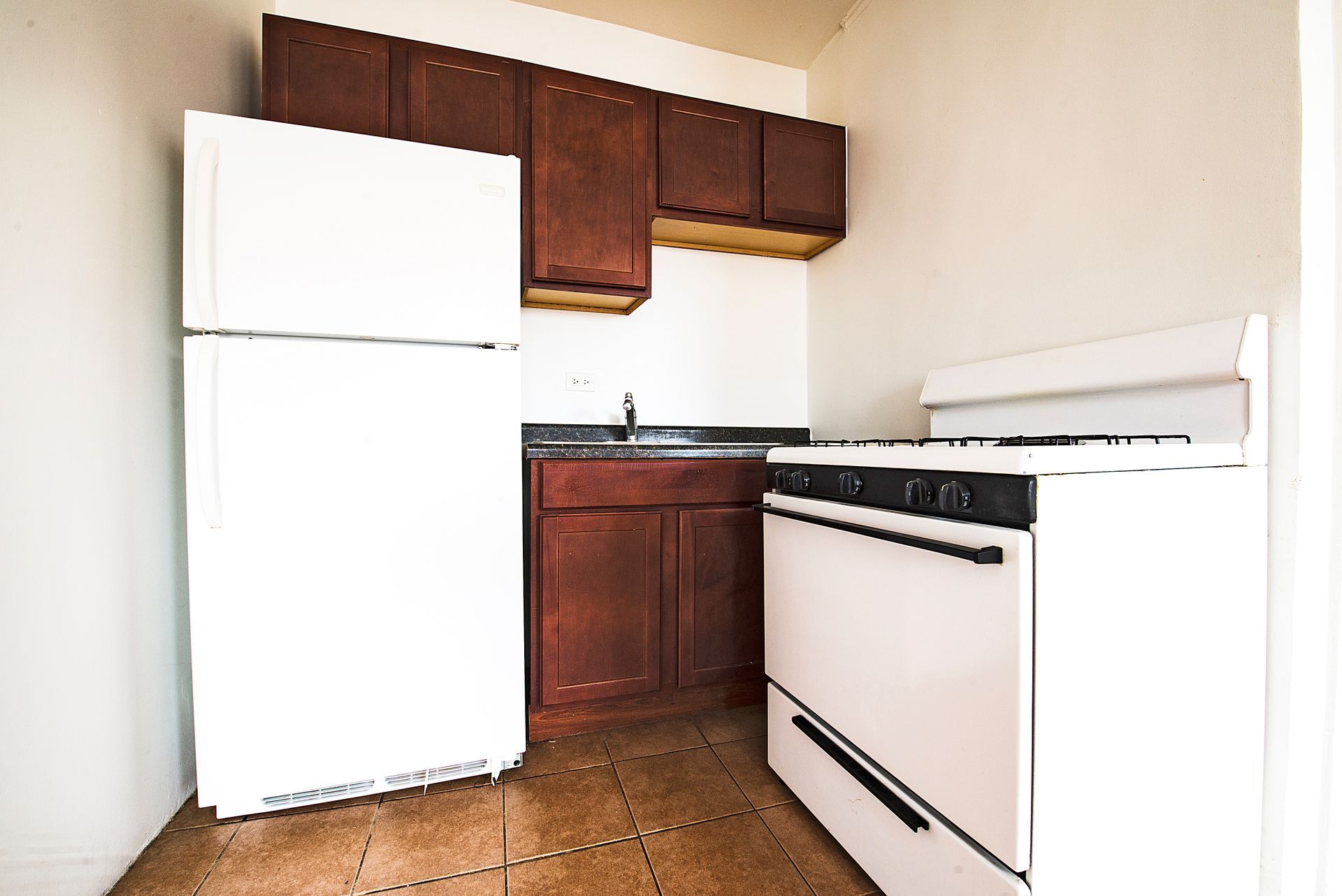 Small kitchen with white refrigerator, stove, and dark wood cabinets. Brown tile floor.