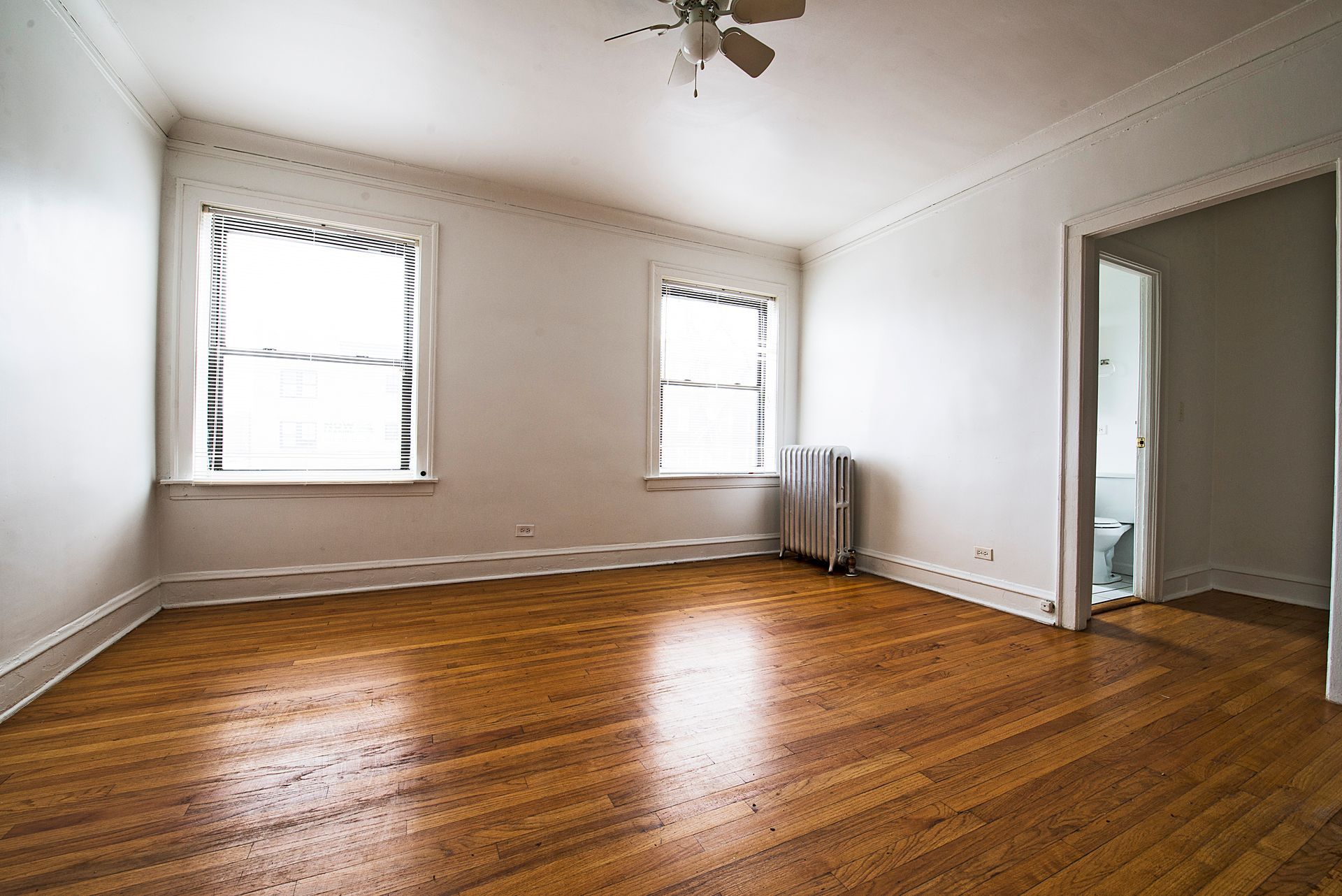 Empty room with hardwood floors, two windows with blinds, radiator, and open doorway.