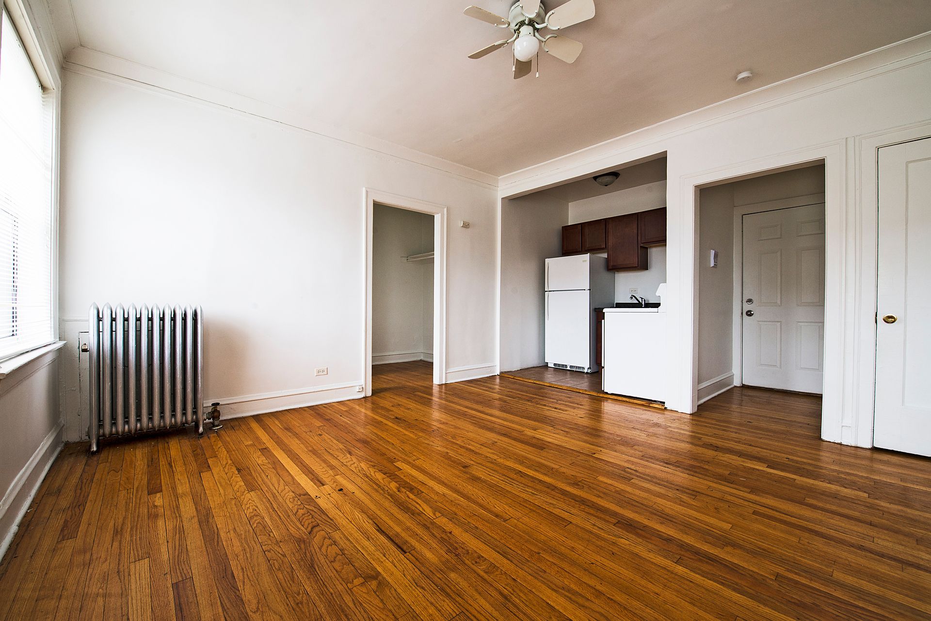 Empty apartment interior with wood floors, radiator, and a small kitchen visible.