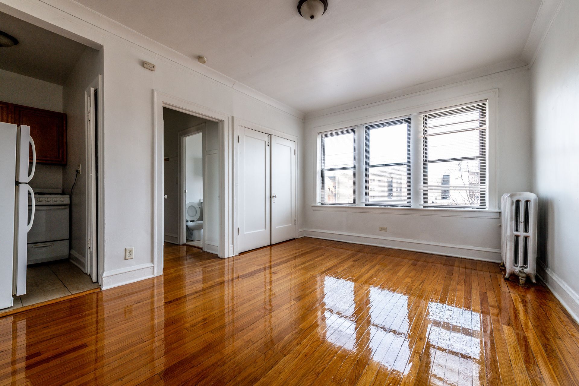Empty apartment interior with wood floors, large windows, and an open kitchen.