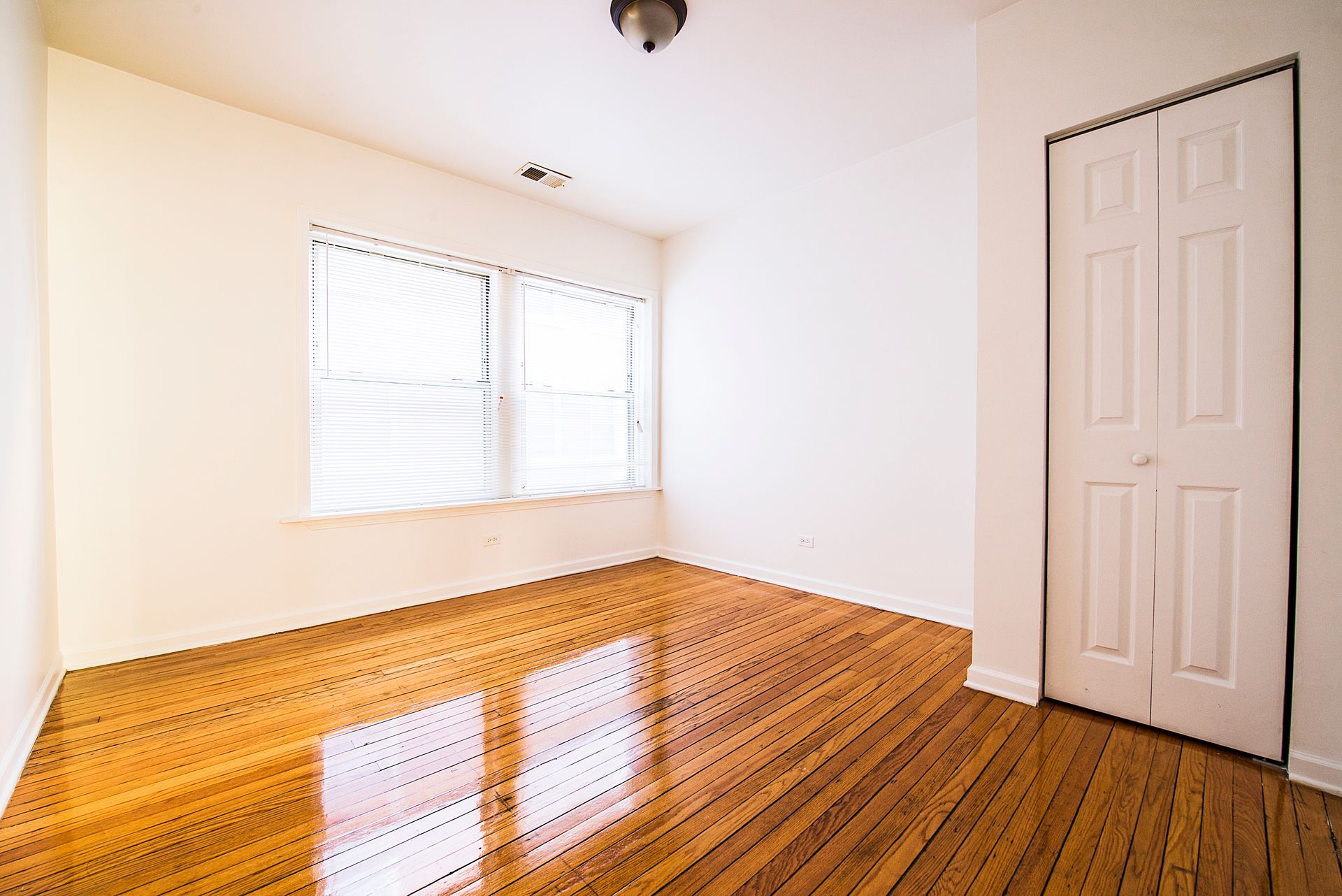 Empty room with hardwood floors, a window with blinds, and a white bi-fold closet door.