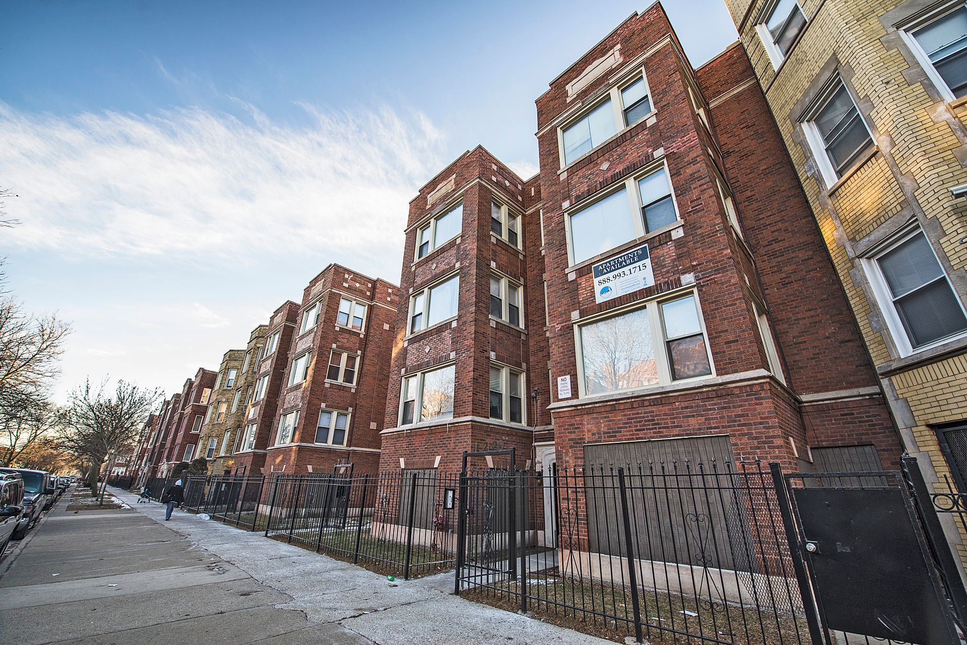 Row of brick apartment buildings with boarded-up windows and a chain-link fence on a city street.