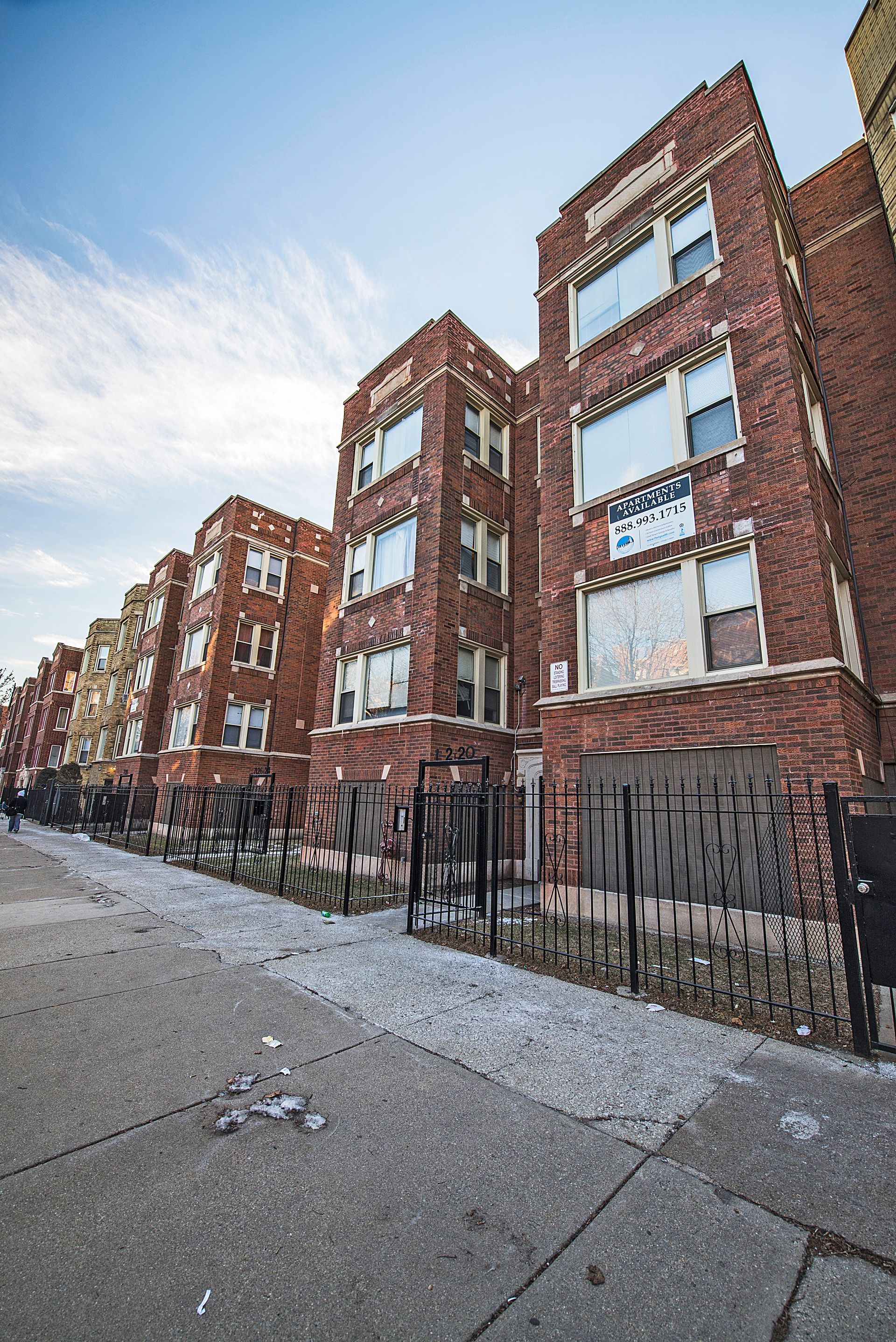 Brick apartment buildings with boarded-up windows and damaged black fence along a sidewalk, daytime.