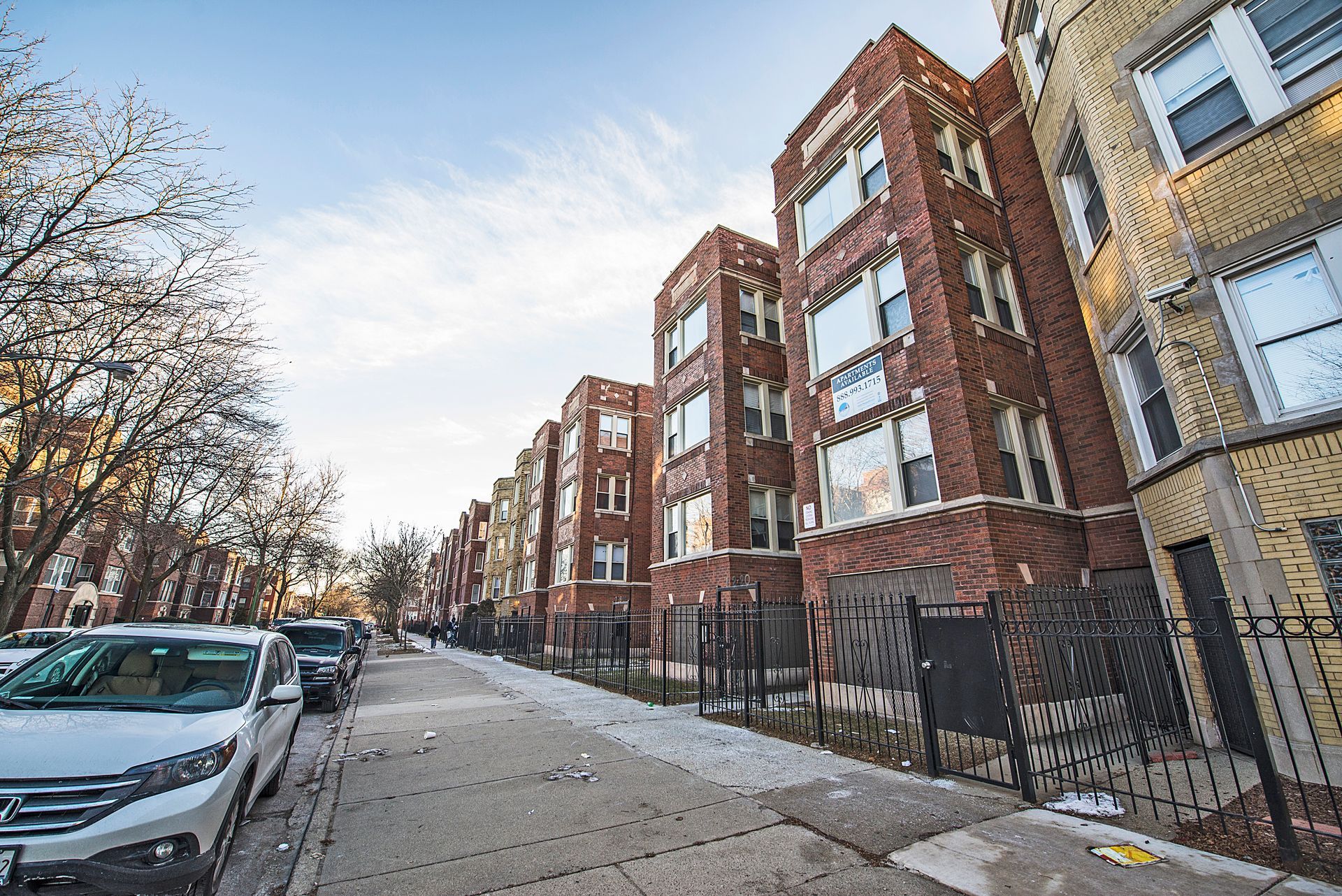 Row of brick apartment buildings on a city street; cars parked along the side, sunny day.