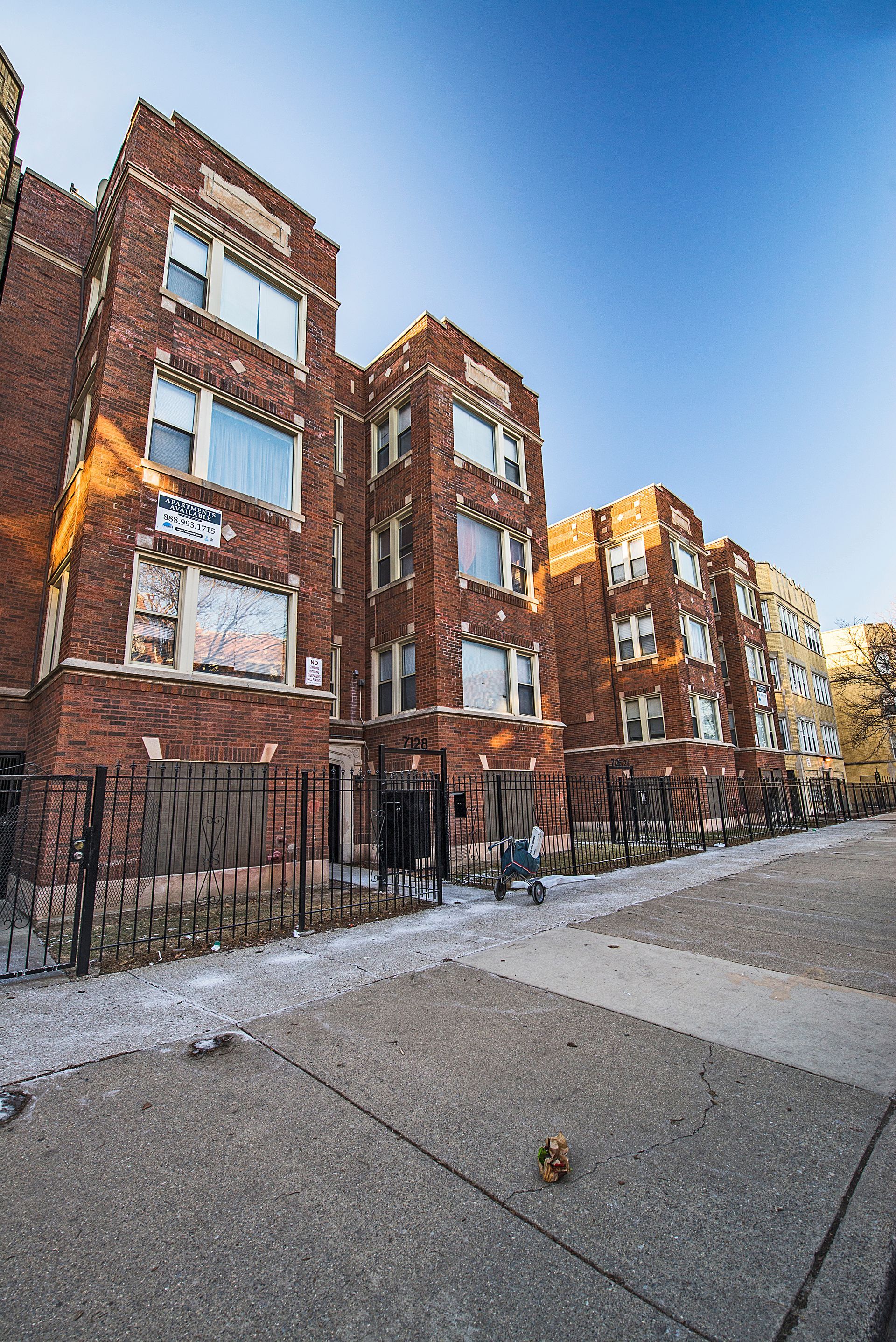 Brick apartment building exterior, front view. Blue sky. Concrete sidewalk. Black fence.