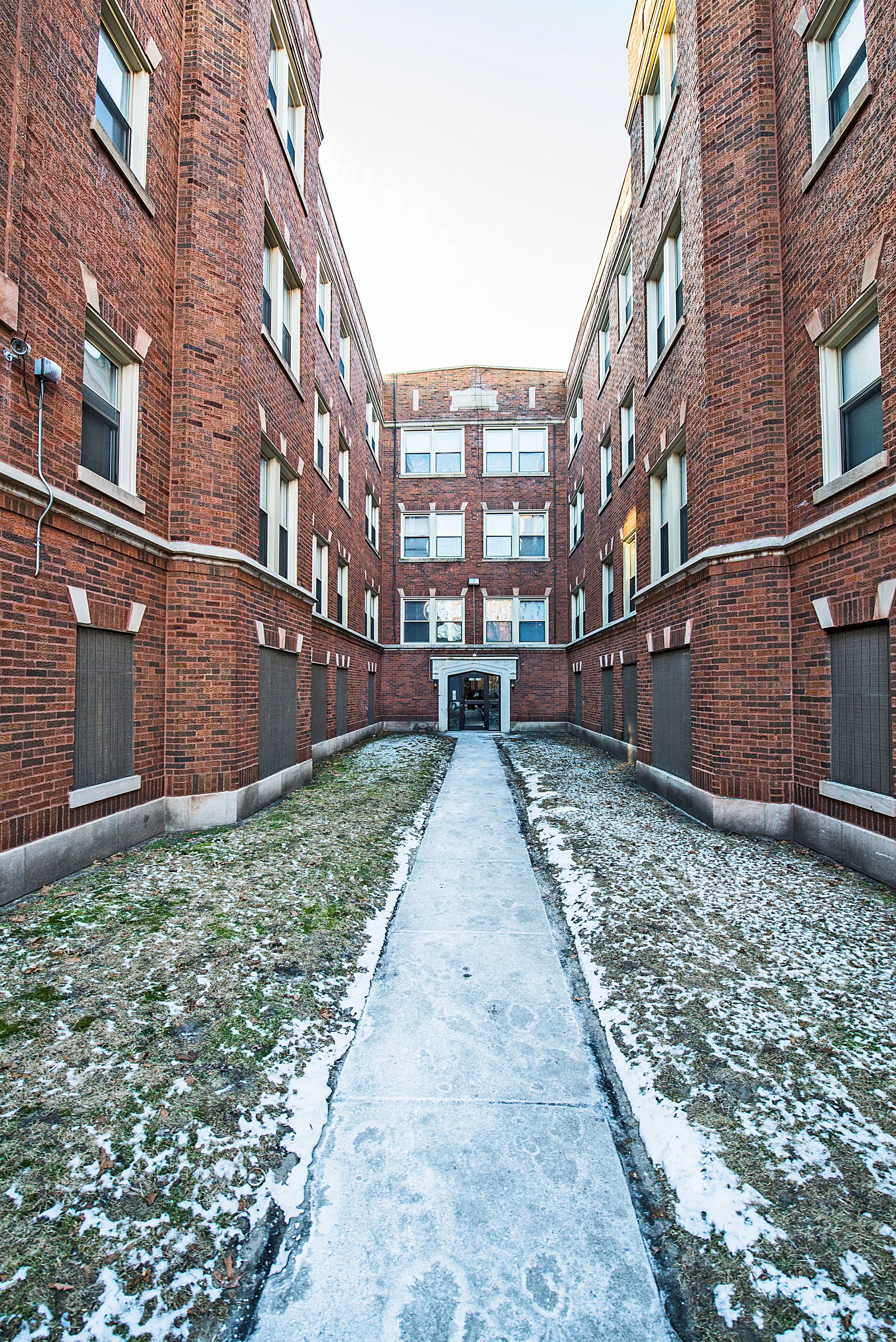 Red brick apartment buildings flank a snow-covered pathway leading to a central entrance.