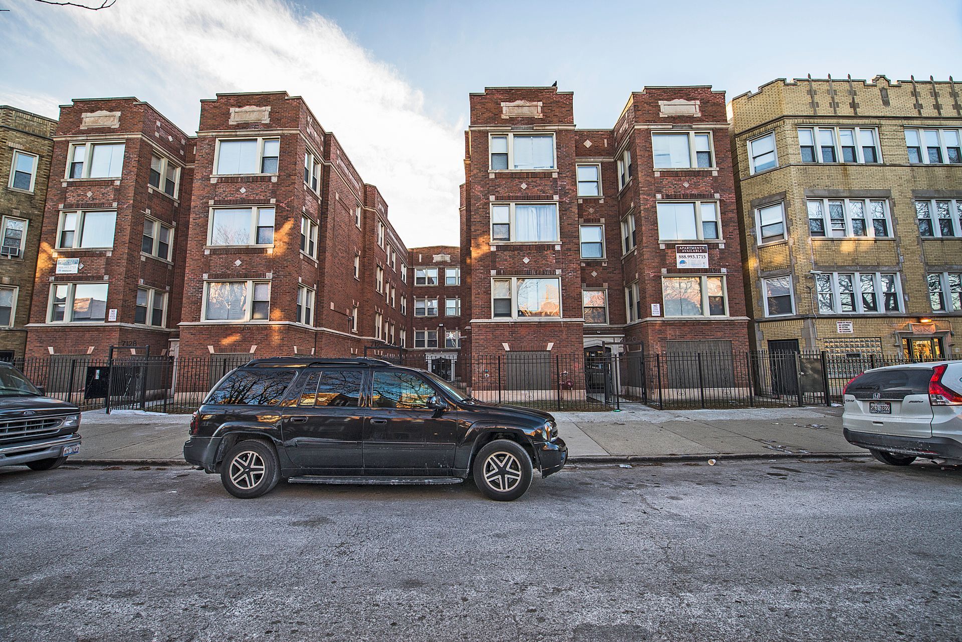 Black SUV parked in front of a brick apartment building with boarded-up windows on a cold day.