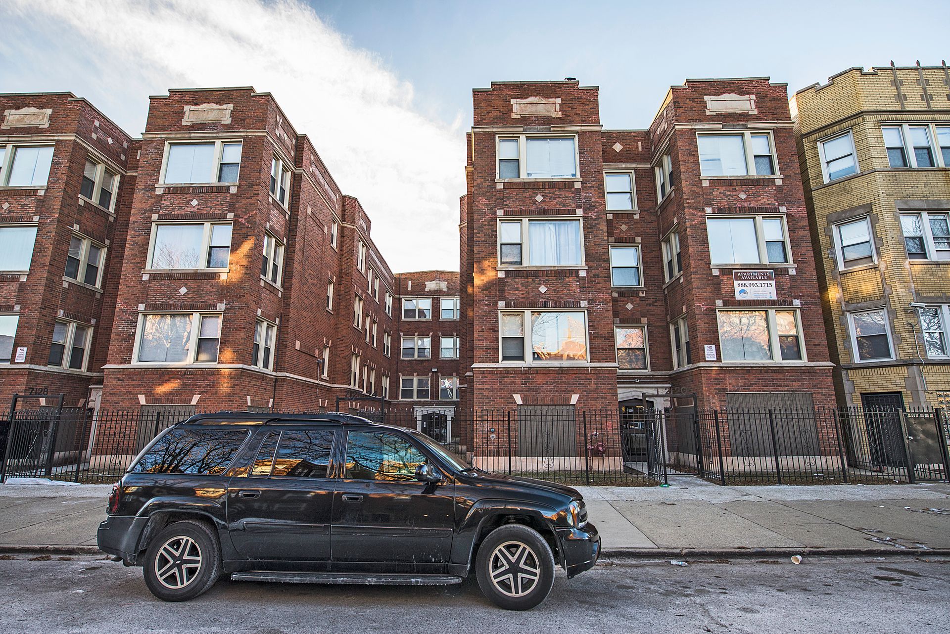 Black SUV parked in front of brick apartment buildings. Person on sidewalk.